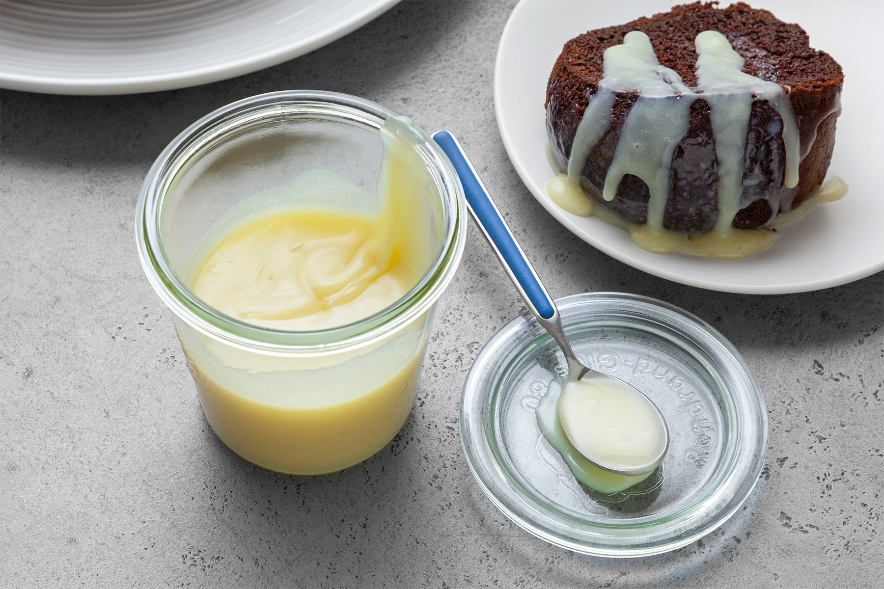 High angle view shot of White Chocolate Sauce; transfer into jar with lid; spoon over cake piece; grey surface;