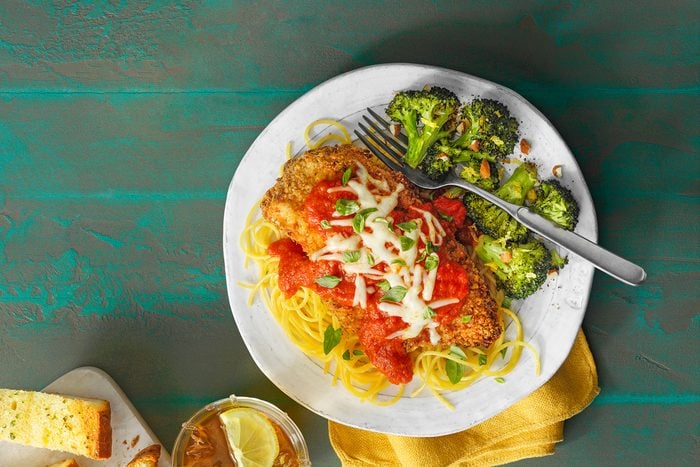 A plate of chicken parmesan topped with tomato sauce and melted cheese sits on a bed of spaghetti. Roasted broccoli is on the side, and a fork rests on the plate. There's a piece of bread and a glass of iced tea with lemon nearby on the table.