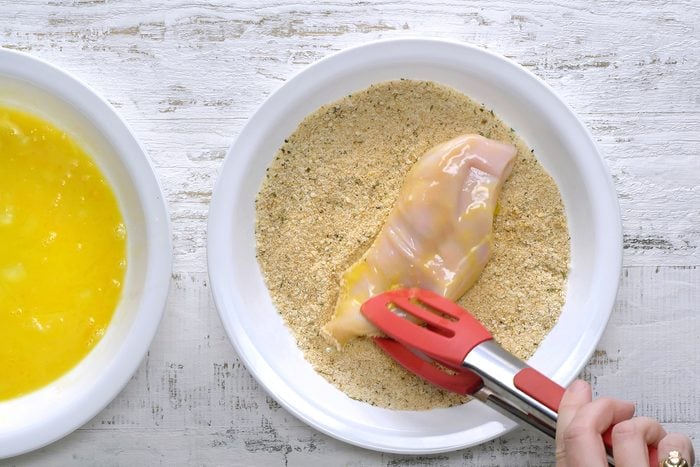 A person uses red tongs to dip a raw chicken breast in breadcrumbs, beside a bowl of beaten eggs on a white wooden surface.
