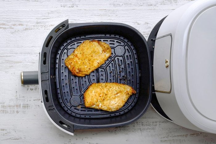 Top view of an air fryer open on a white wooden surface, featuring two cooked, seasoned chicken breasts inside the fryer basket.