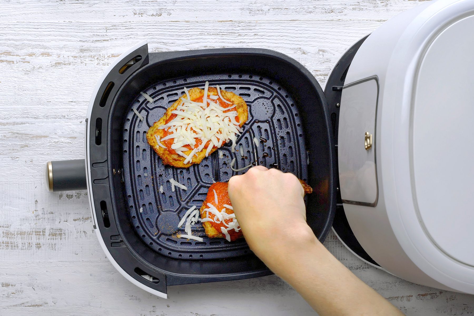 A hand placing a piece of breaded chicken topped with shredded cheese into an air fryer. Another piece of chicken is already inside. The air fryer is white and black, resting on a light wooden surface.