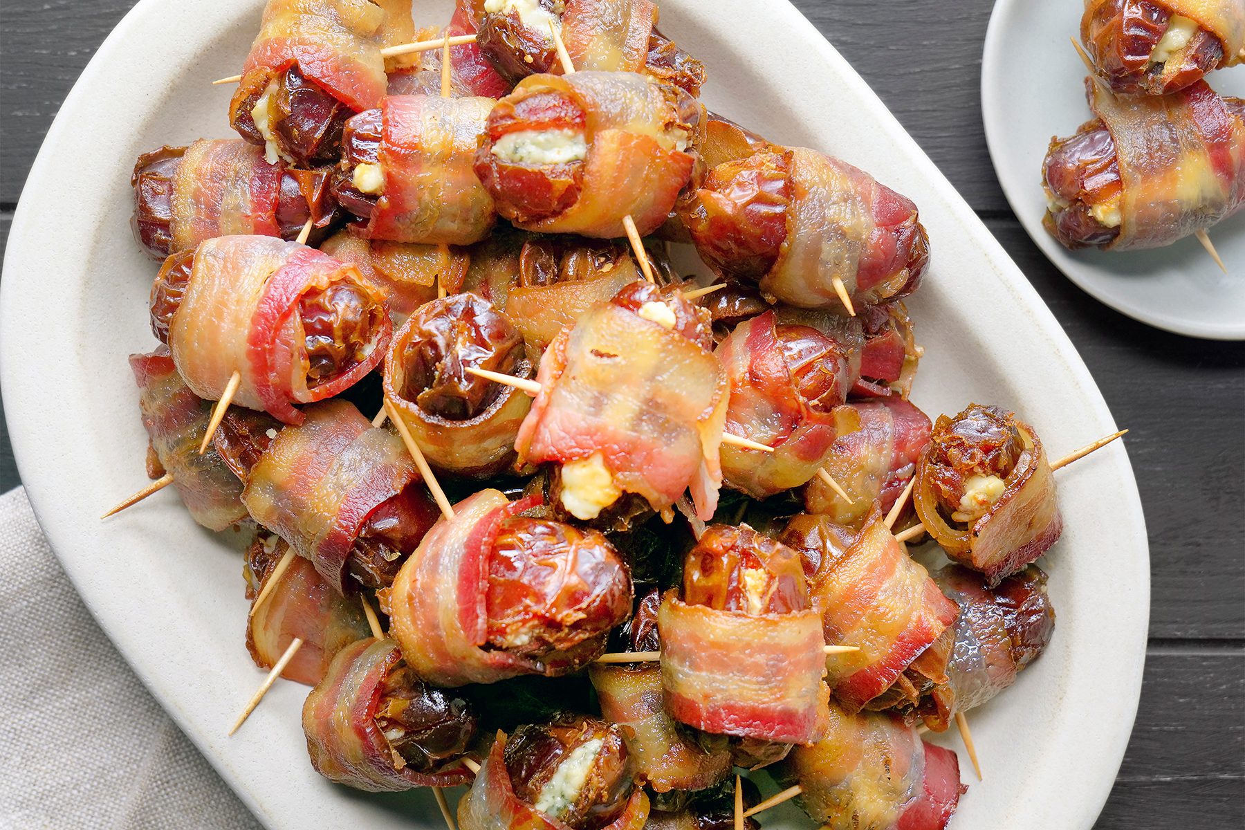 A white plate filled with bacon-wrapped dates, each secured with toothpicks. The dates appear caramelized and arranged neatly, showcasing the crispy bacon exterior. A smaller plate with more dates is partially visible in the background.