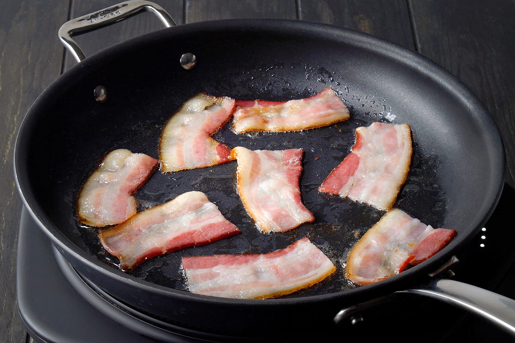 Slices of bacon sizzling in a black frying pan on a stovetop, with some pieces turning golden brown. The bacon lies in its own grease, indicating they are in the process of cooking. The background is dark, highlighting the pan's contents.
