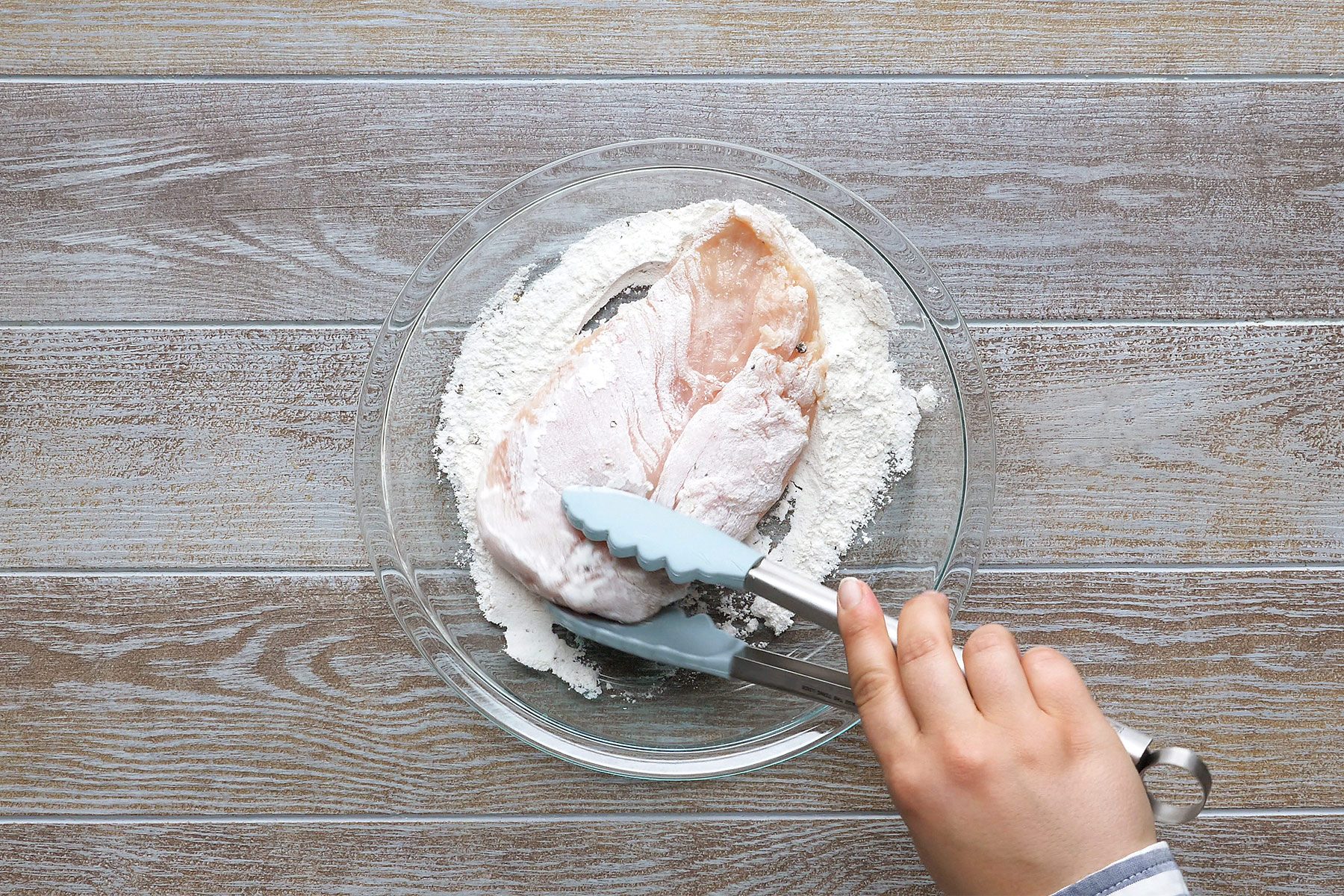 overhead shot of a person using tongs to dredge a chicken breast in flour; the chicken breast is in a glass bowl with flour and the person is using the tongs to turn the chicken breast in the flour; the background is a light brown woodgrain