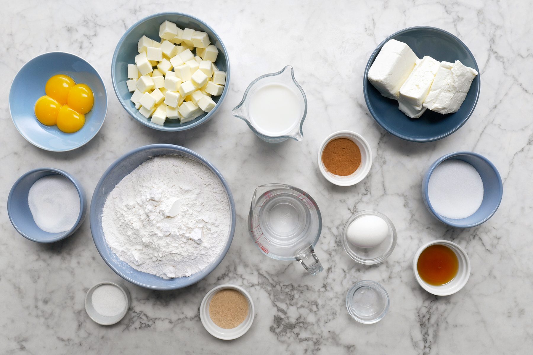Ingredients for a baking recipe arranged on a marble surface, including flour, sugar, butter, milk, cream cheese, egg yolks, a whole egg, vanilla extract, cinnamon, and yeast, each in individual bowls or containers.