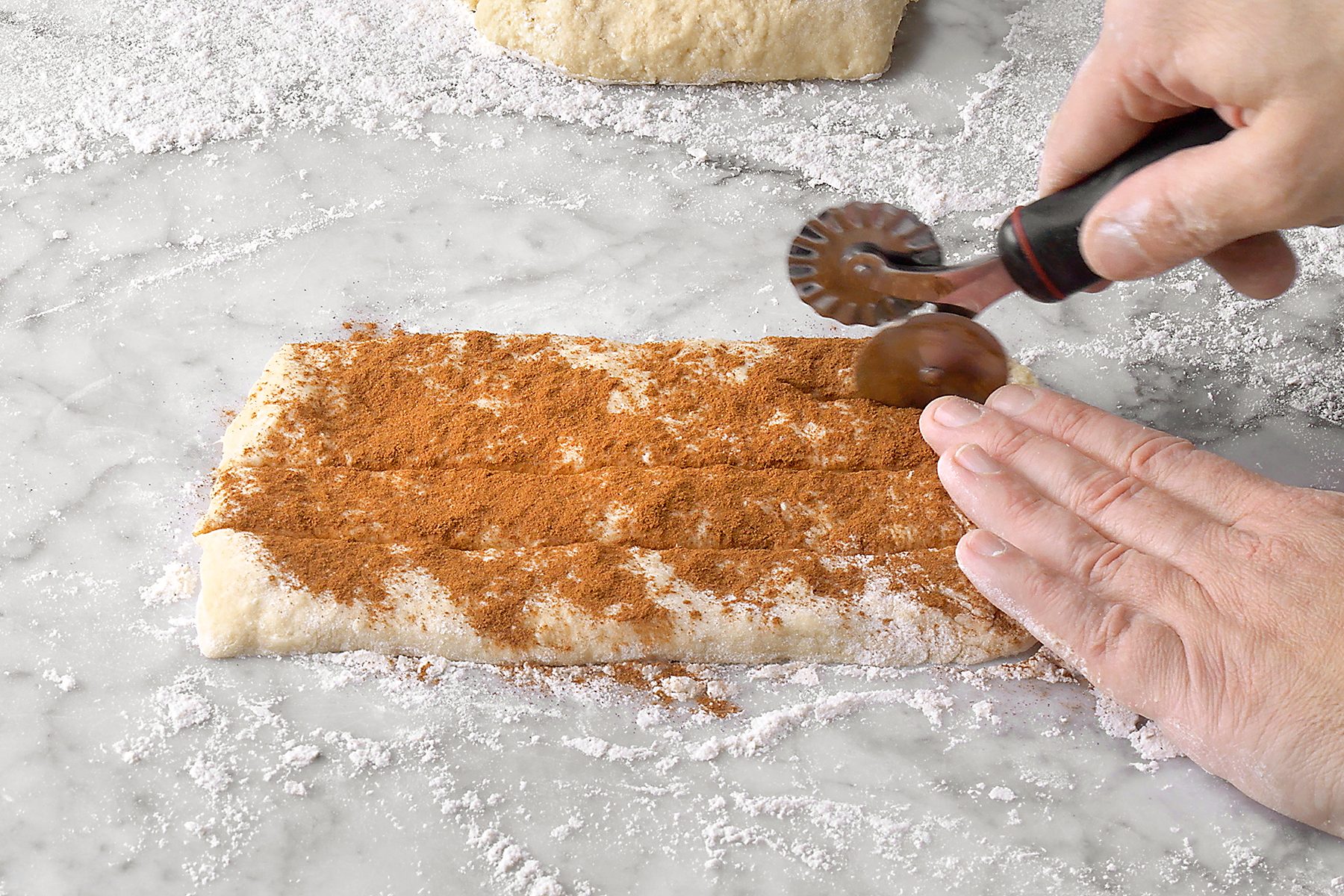 Hands are rolling dough with cinnamon on a floured surface using a pastry cutter.