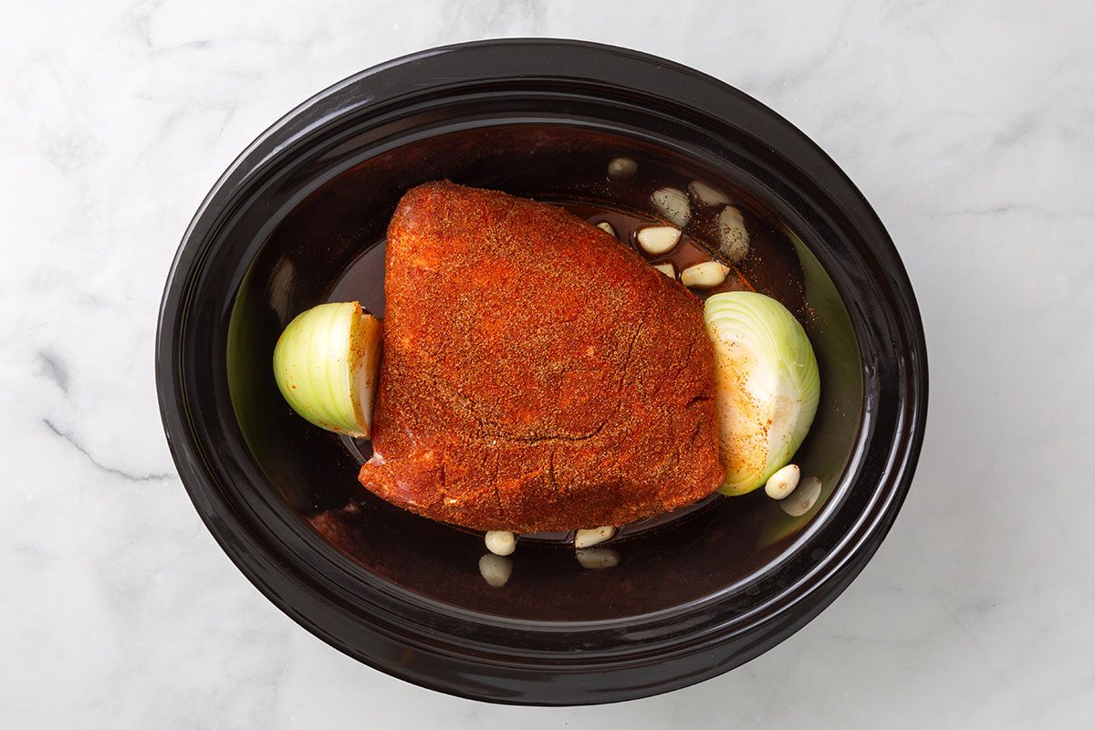 Pork shoulder covered in ground chipotle, cumin and salt rub in a slow cooker with a halved onion and garlic cloves for step one of Chipotle Carnitas recipe for Taste of Home