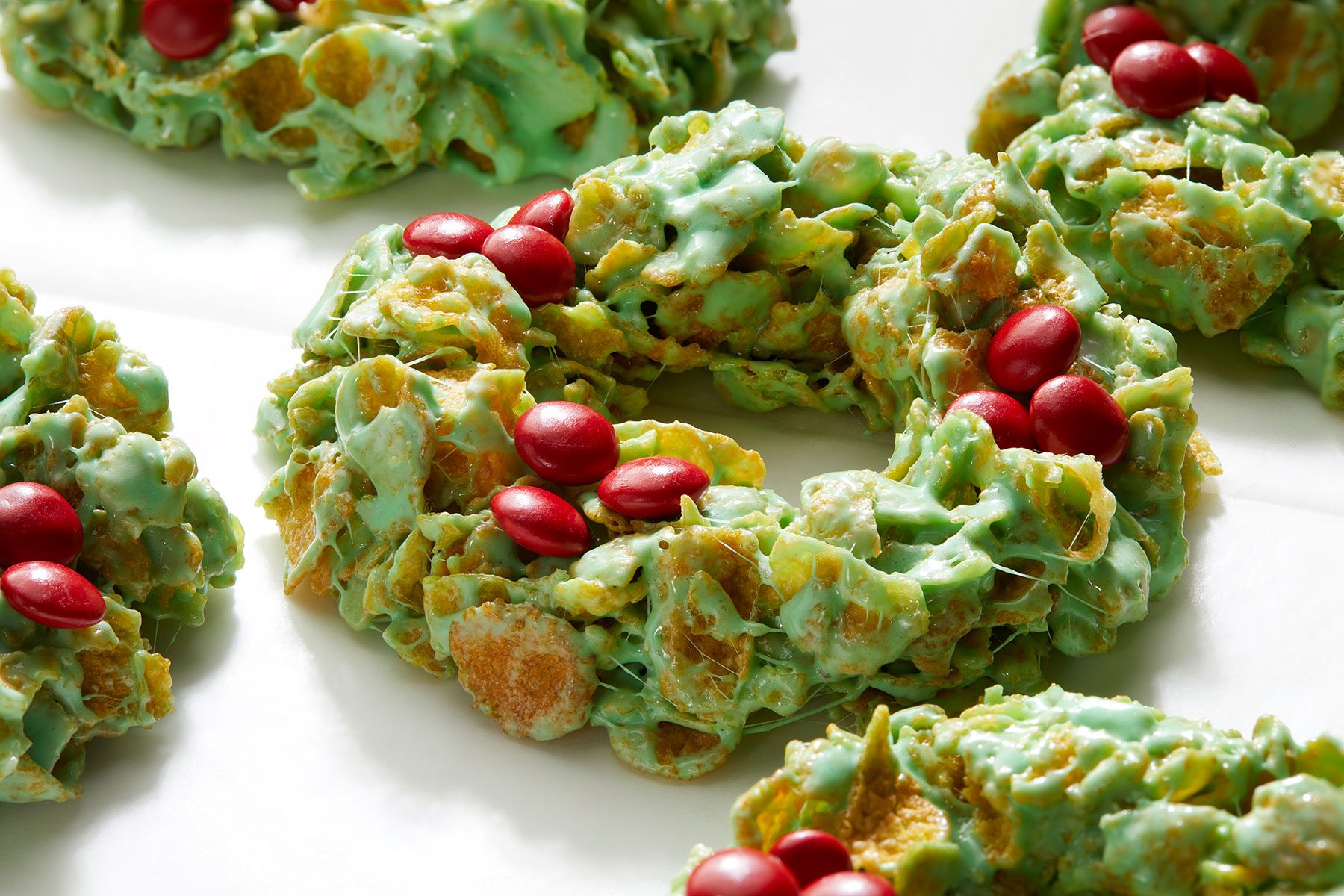 Festive green wreath-shaped cookies made of cornflakes, topped with clusters of red candy pieces, placed on a white background.