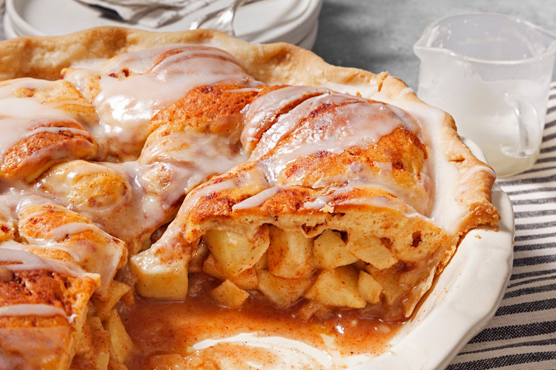 3/4th shot of a white pie dish holds an apple pie, its golden-brown crust glistening under a drizzle of creamy glaze; The pie is sliced open, revealing a generous filling of warm, spiced apples; there is a small white plate and a glass measuring cup filled with a white liquid in the background ; the pie dish is sitting on a white and grey striped linen napkin