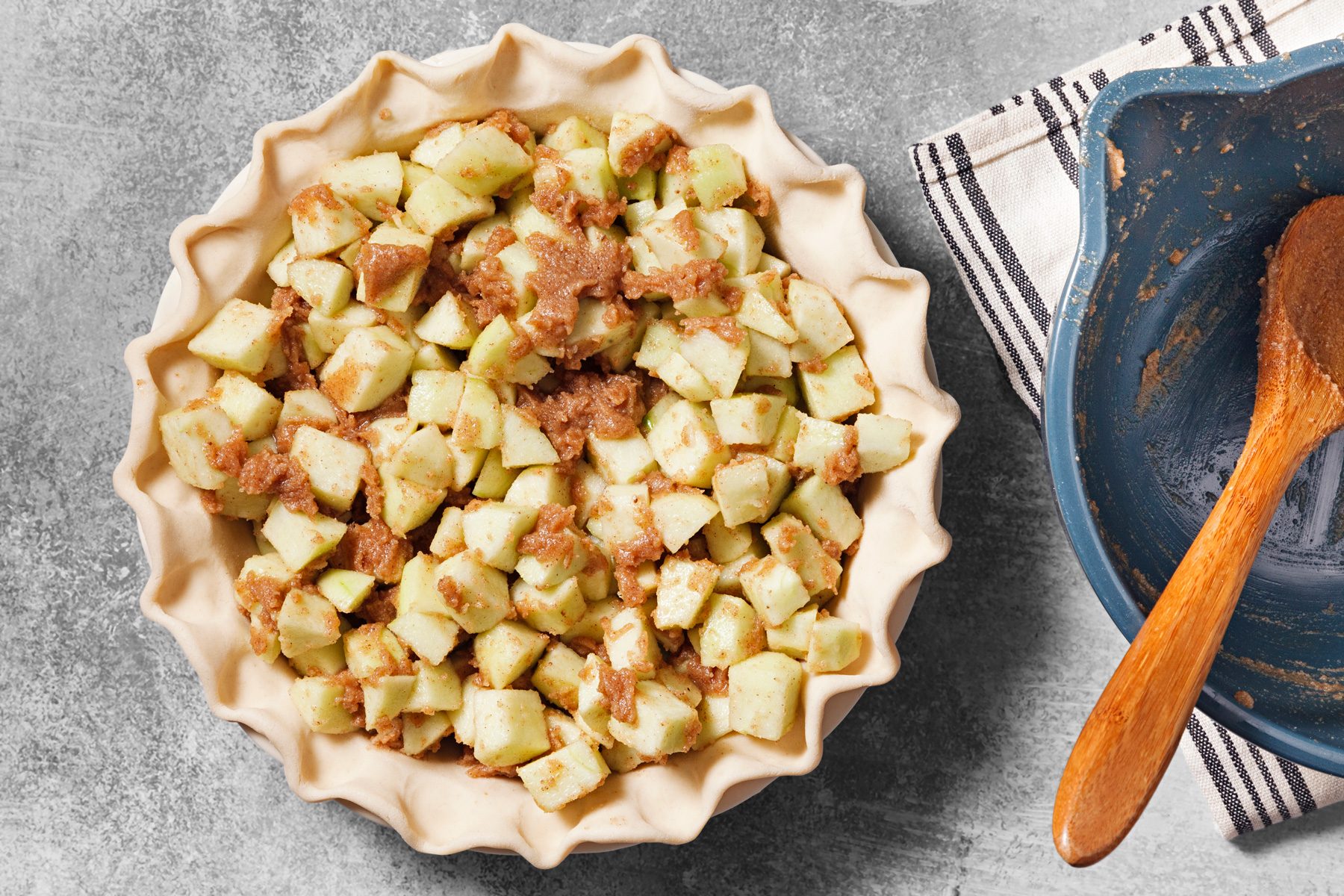 overhead shot of a close-up of a pie crust filled with diced apples and a brown sugar, cinnamon mixture; the crust is in a pie pan and the filling is almost level with the top of the crust; a wooden spoon lies next to a blue bowl which is partially filled with the remainder of the apple filling and is on top of a striped kitchen towel