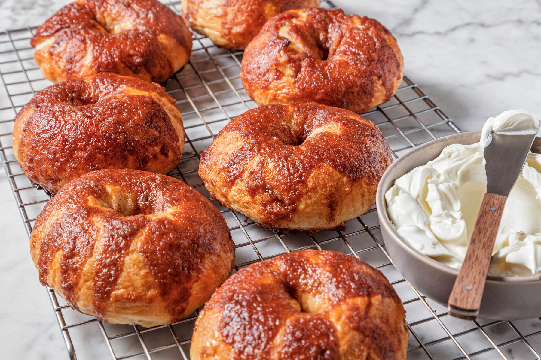 Close shot of Copycat Panera Bread Cinnamon Crunch Bagels being removed from pans and placed on wire racks to cool, with a bowl of whipped cream and a knife on a marble surface.