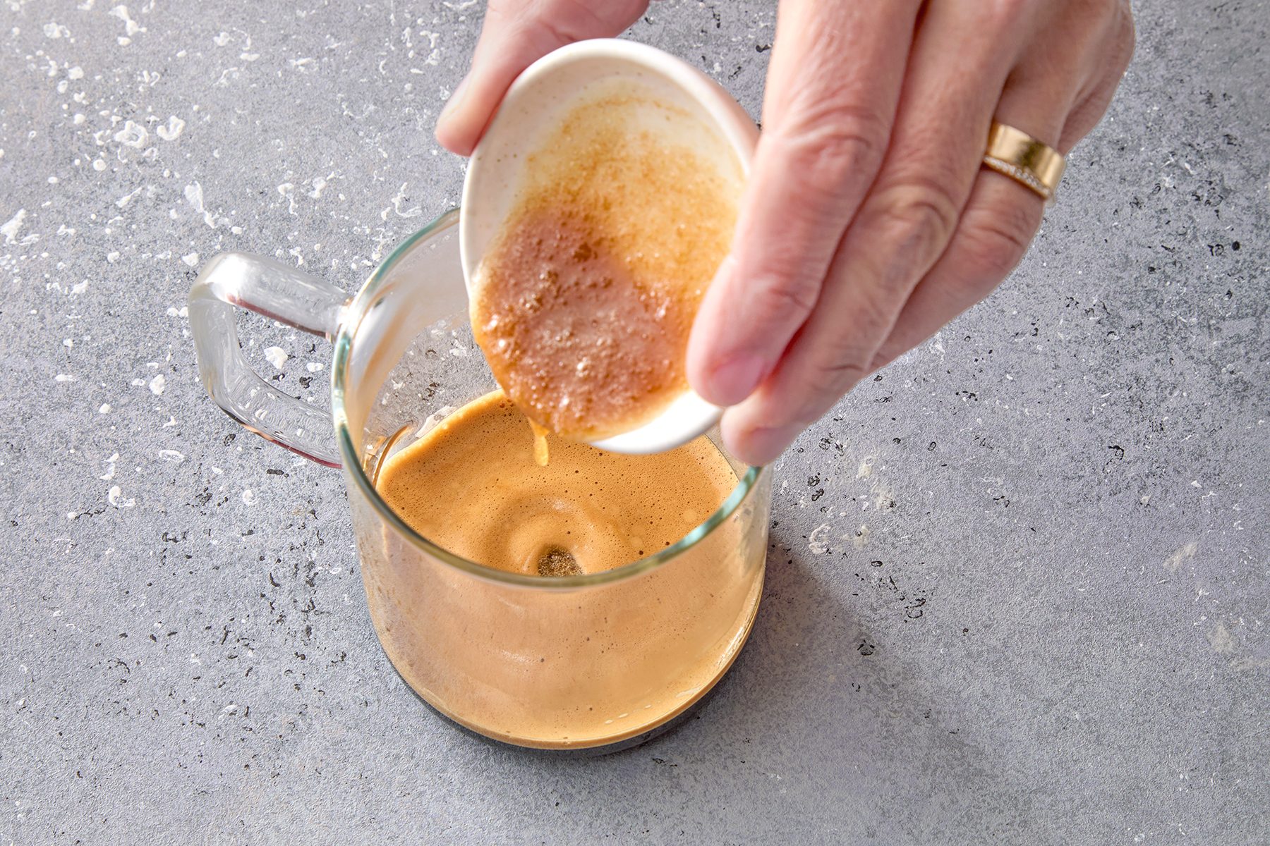 A person's hand pouring coffee froth from a small white dish into a glass mug of creamy coffee on a textured gray surface.