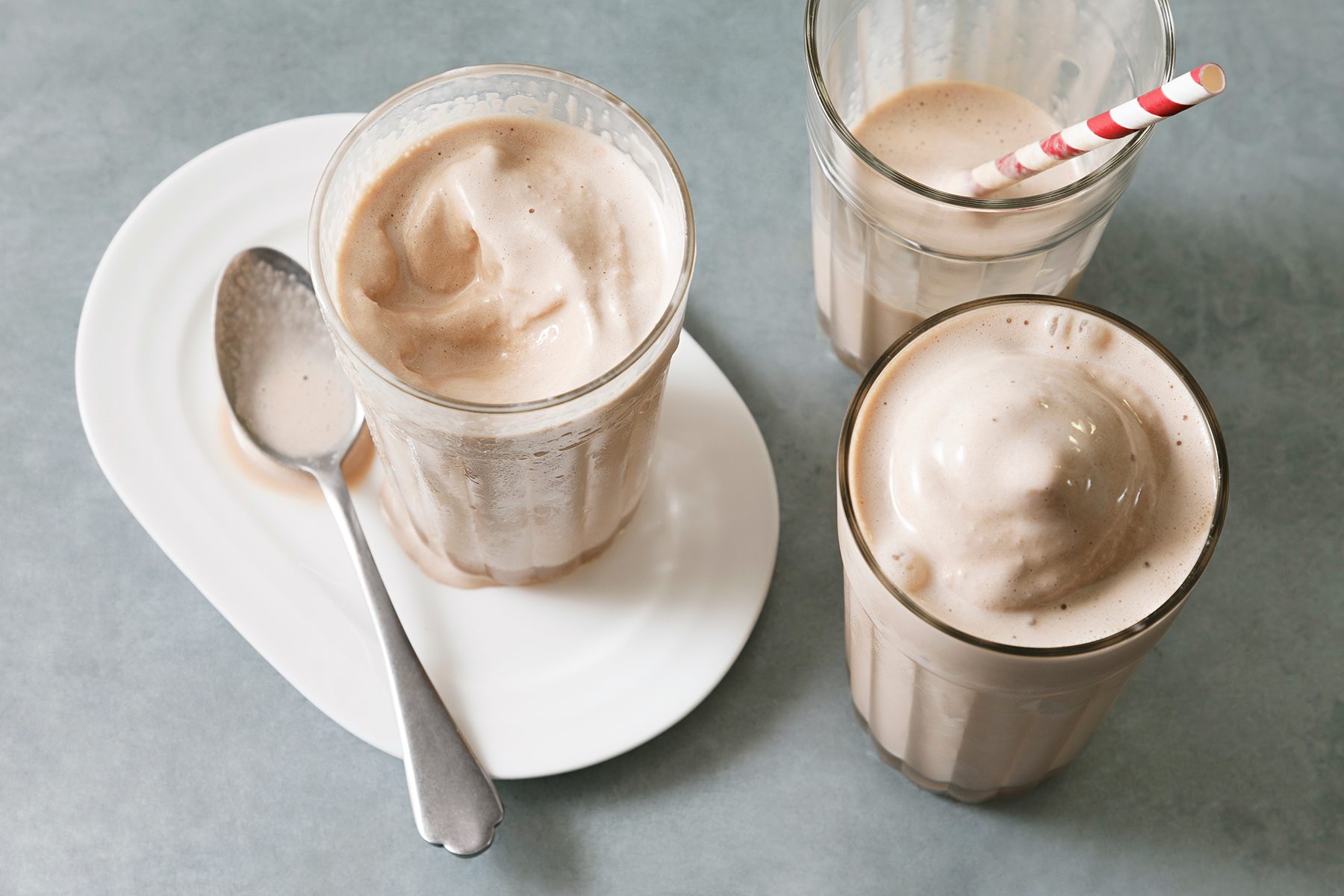 Three glasses of chocolate milkshake on a gray surface, one with a red and white striped straw. A spoon rests on a white plate beside the drinks.