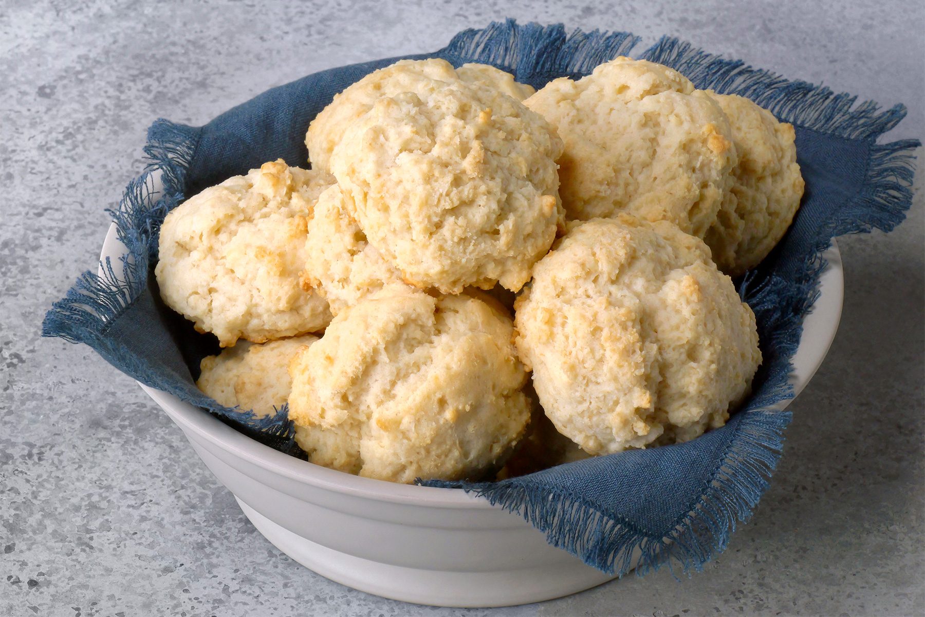 A white bowl filled with golden-brown biscuits sits on a blue cloth. The biscuits have a rough, textured surface and are arranged in a cozy stack. The bowl is placed on a lightly speckled gray surface.