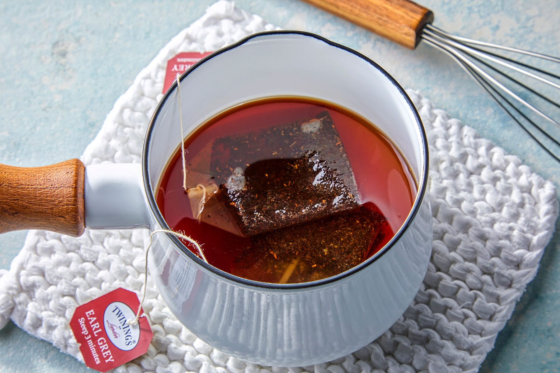A white mug filled with Earl Grey tea steeps a tea bag on a textured white mat. Two tea bag tags labeled "Twinings Earl Grey" are visible, with a whisk nearby on a light blue surface.