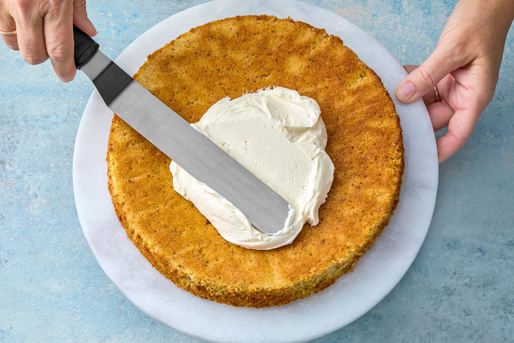 A person spreading frosting onto a round cake with a spatula. The cake is placed on a white marble surface against a light blue background.
