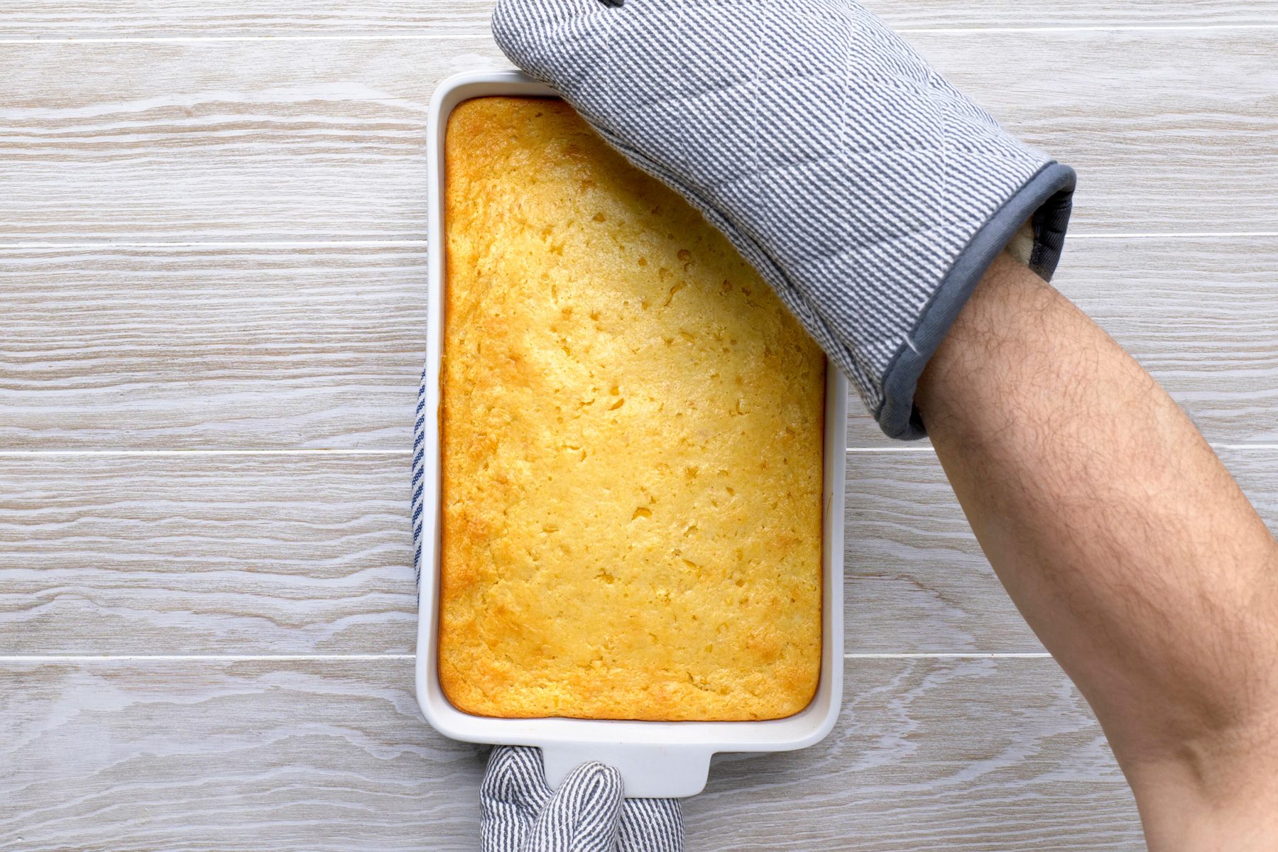 overhead shot of a top-down view of a baking dish containing a golden yellow cornbread; the baking dish is on a white wooden countertop; a hand wearing a striped oven mitt is reaching in to grab the handle of the baking dish;