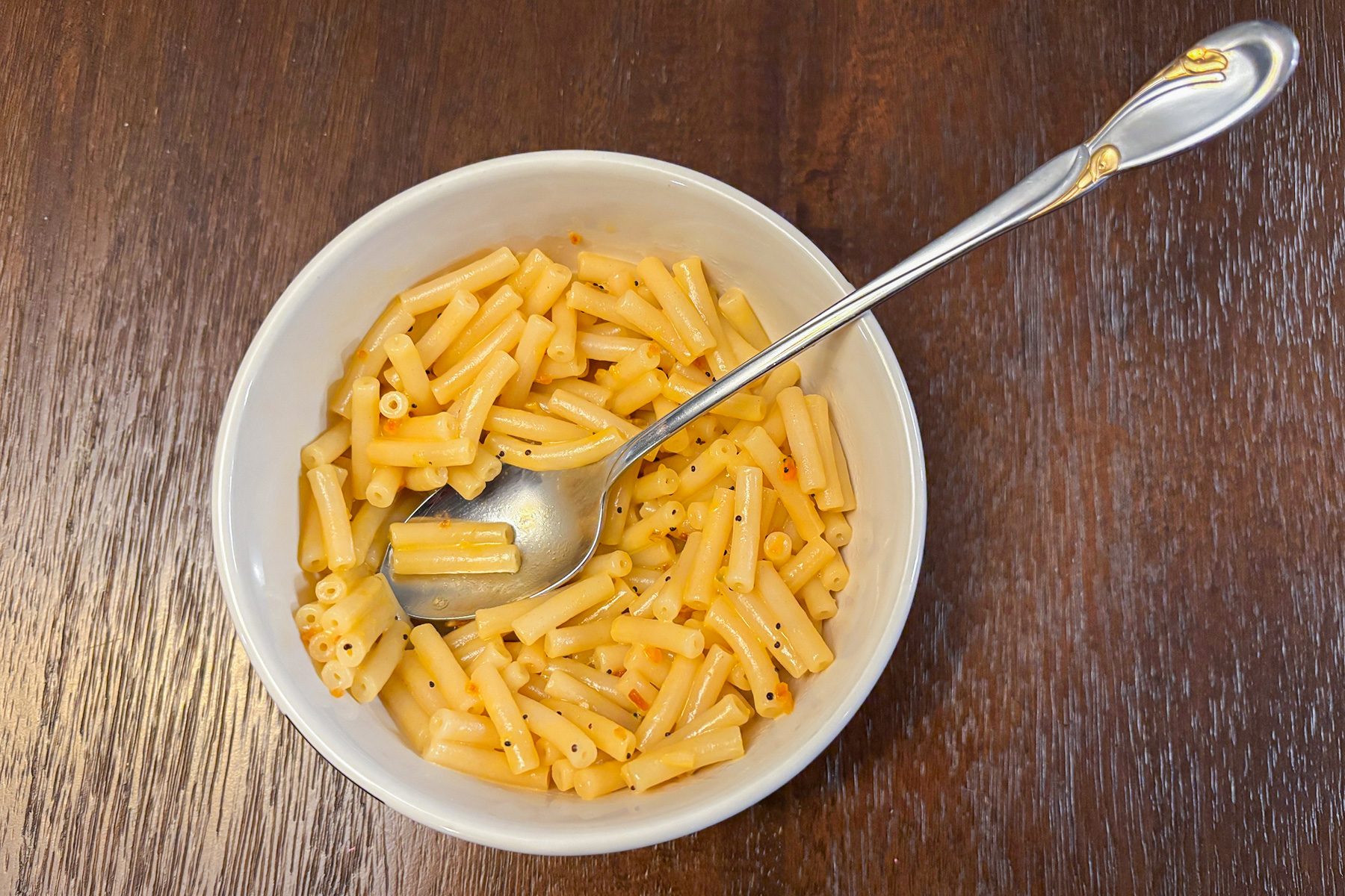 A white bowl filled with cooked macaroni pasta on a wooden table. A silver spoon is resting in the bowl. The macaroni is lightly coated with a yellowish sauce.