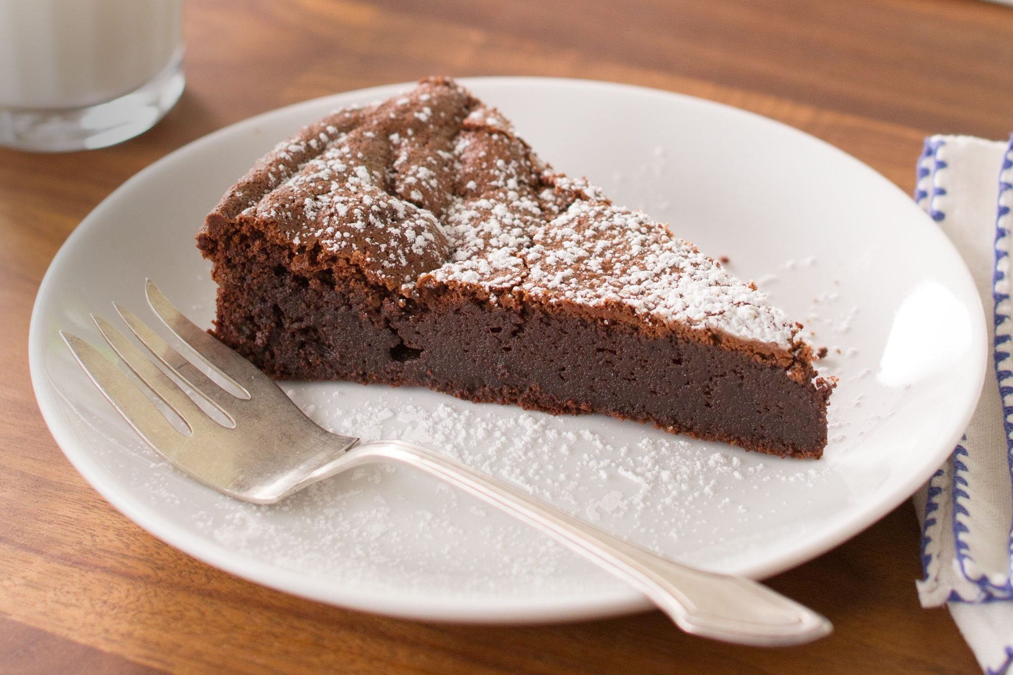 A slice of Flourless Chocolate Torte dusted with powdered sugar sits on a white plate with a fork beside it. The plate is placed on a wooden table next to a blue-striped napkin.