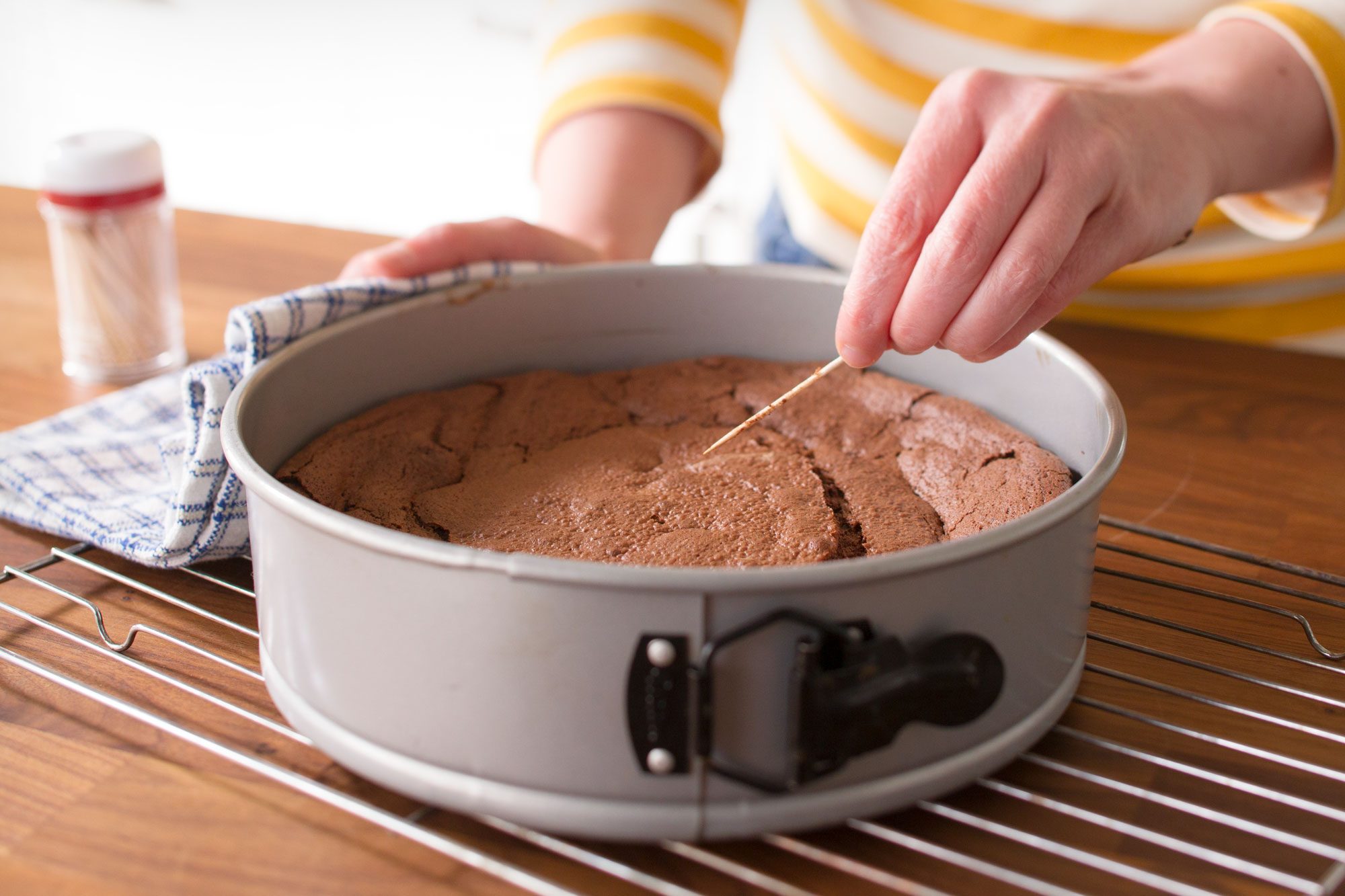 Person testing the doneness of a chocolate cake with a toothpick in a round baking pan on a cooling rack.