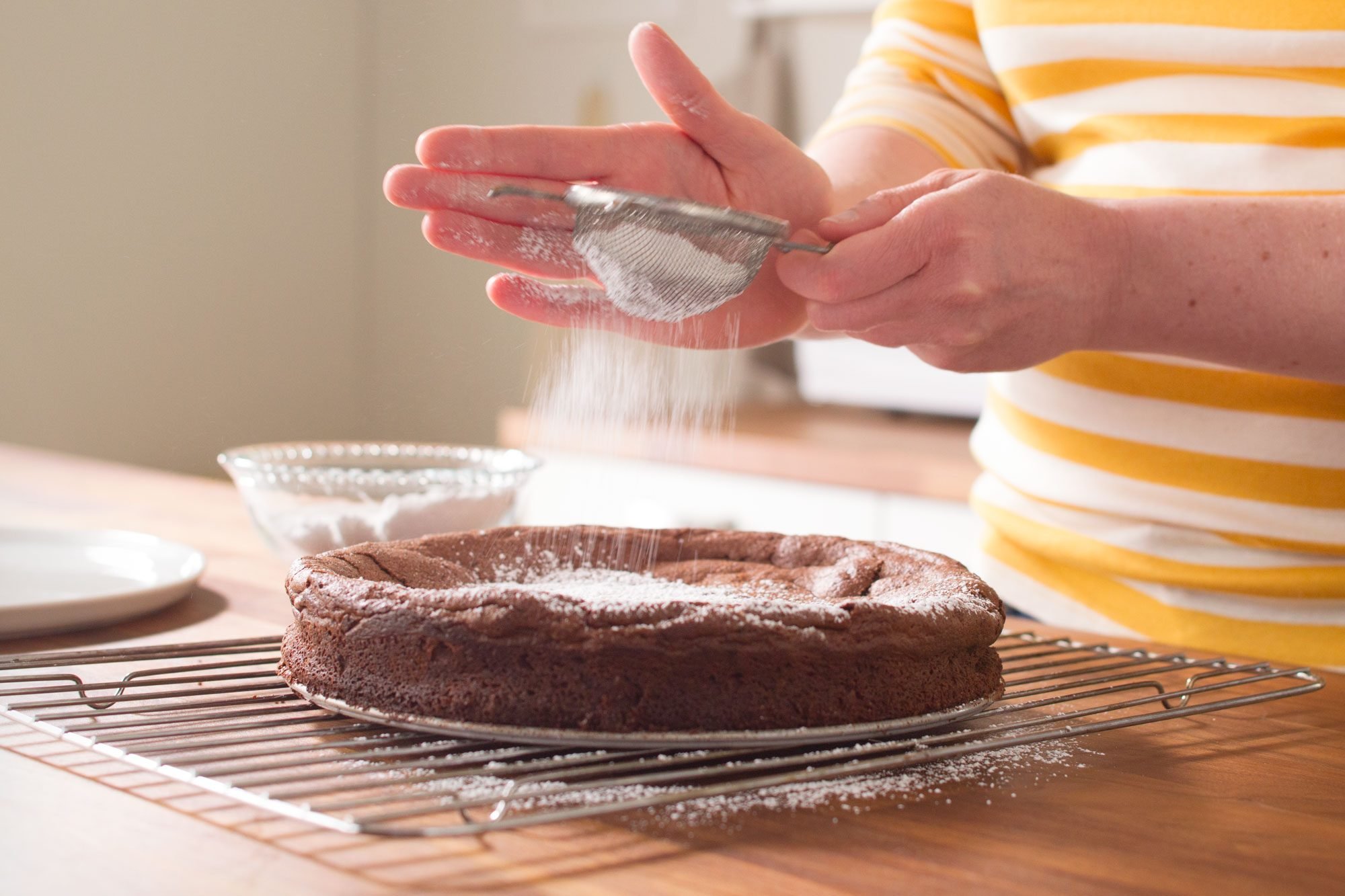 Person dusting powdered sugar onto a chocolate cake using a sieve. The cake rests on a cooling rack on a wooden countertop. The person is wearing a yellow and white striped shirt.