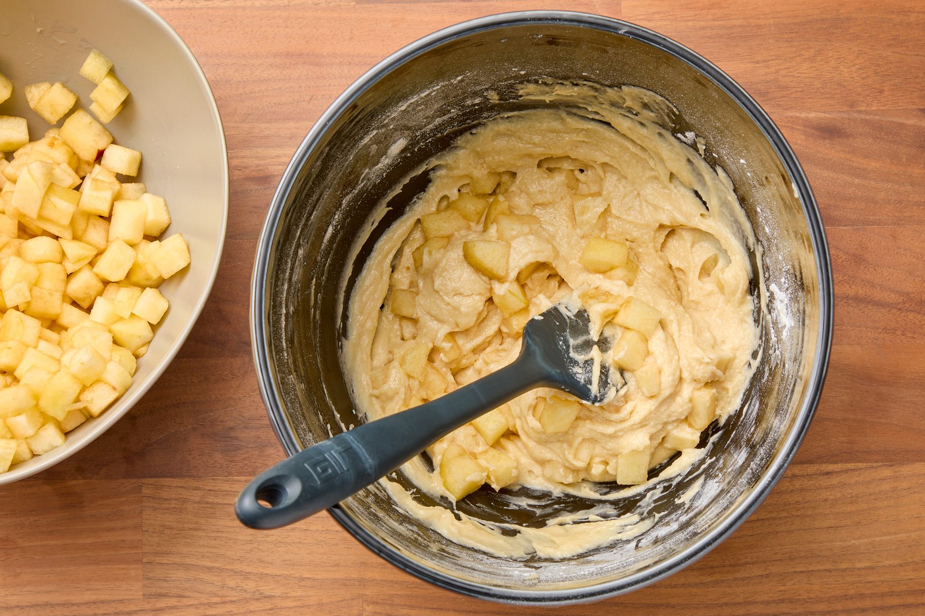 Overhead shot of a black bowl filled with creamy batter containing diced apples, with a black rubber spatula inside. A grey bowl of diced apples is positioned to the left. Both bowls are placed on a wooden surface.