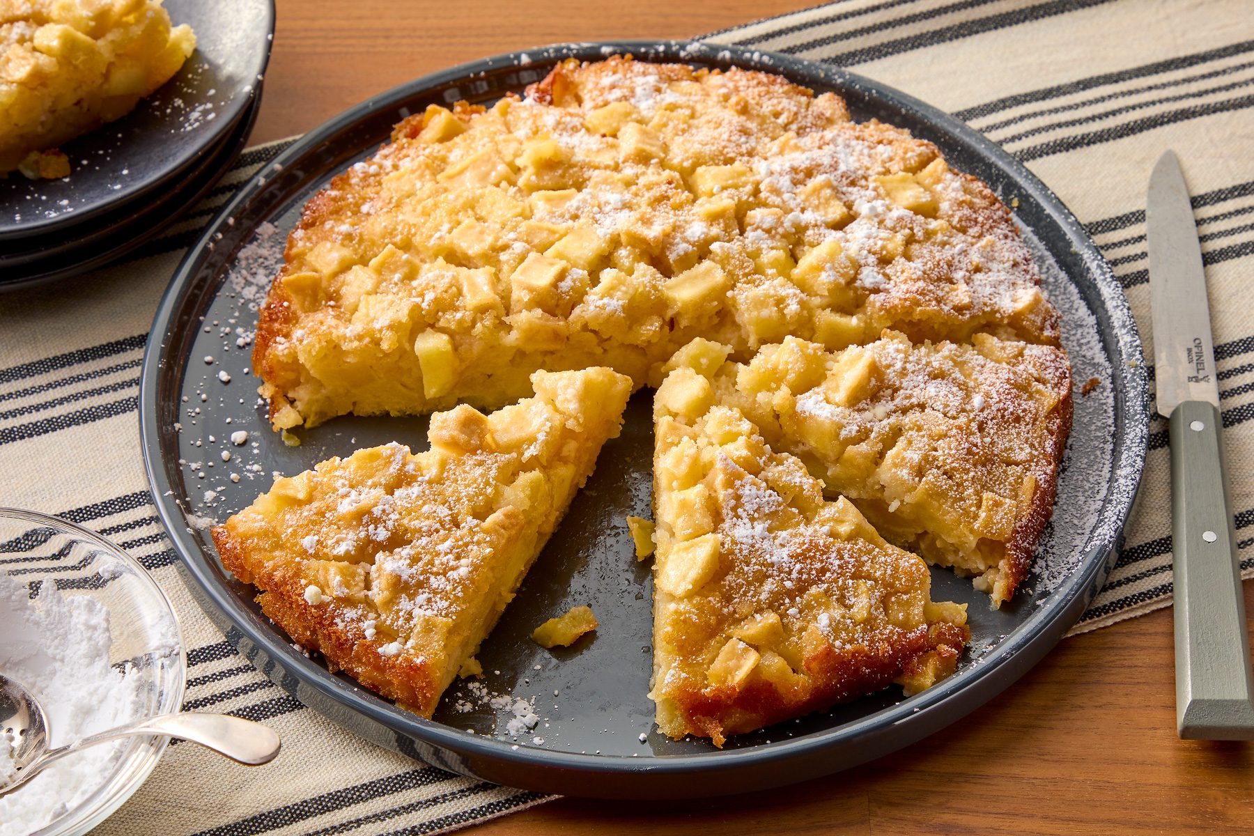 Three-quarter shot of an apple cake with a dusting of powdered sugar, sitting on a gray plate with four slices missing. A knife is placed on the right side of the plate, and a bowl of powdered sugar sits on the left. The plate rests on a brown table with a striped beige and black placemat underneath.