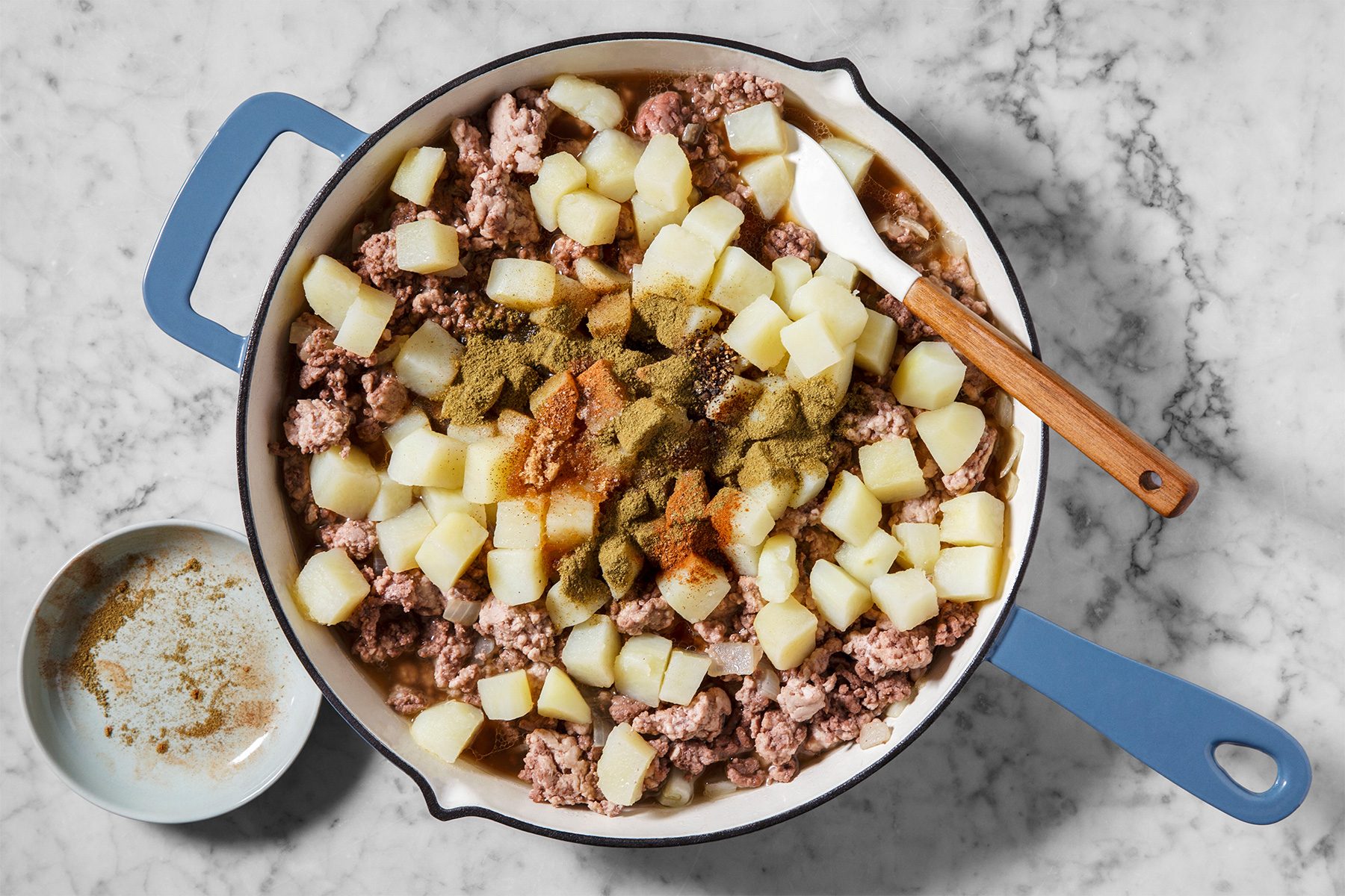 A blue-handled pan on a marble surface contains diced potatoes and ground beef with a sprinkle of spices. A wooden-handled spatula rests on the pan's edge. A small bowl with spices is next to the pan.