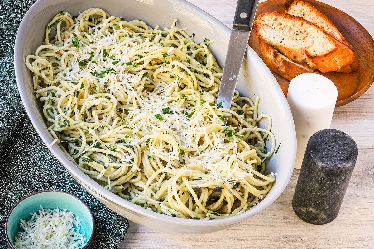 A serving bowl of garlic spaghetti, with extra Parmesan and baguette slices on the side.