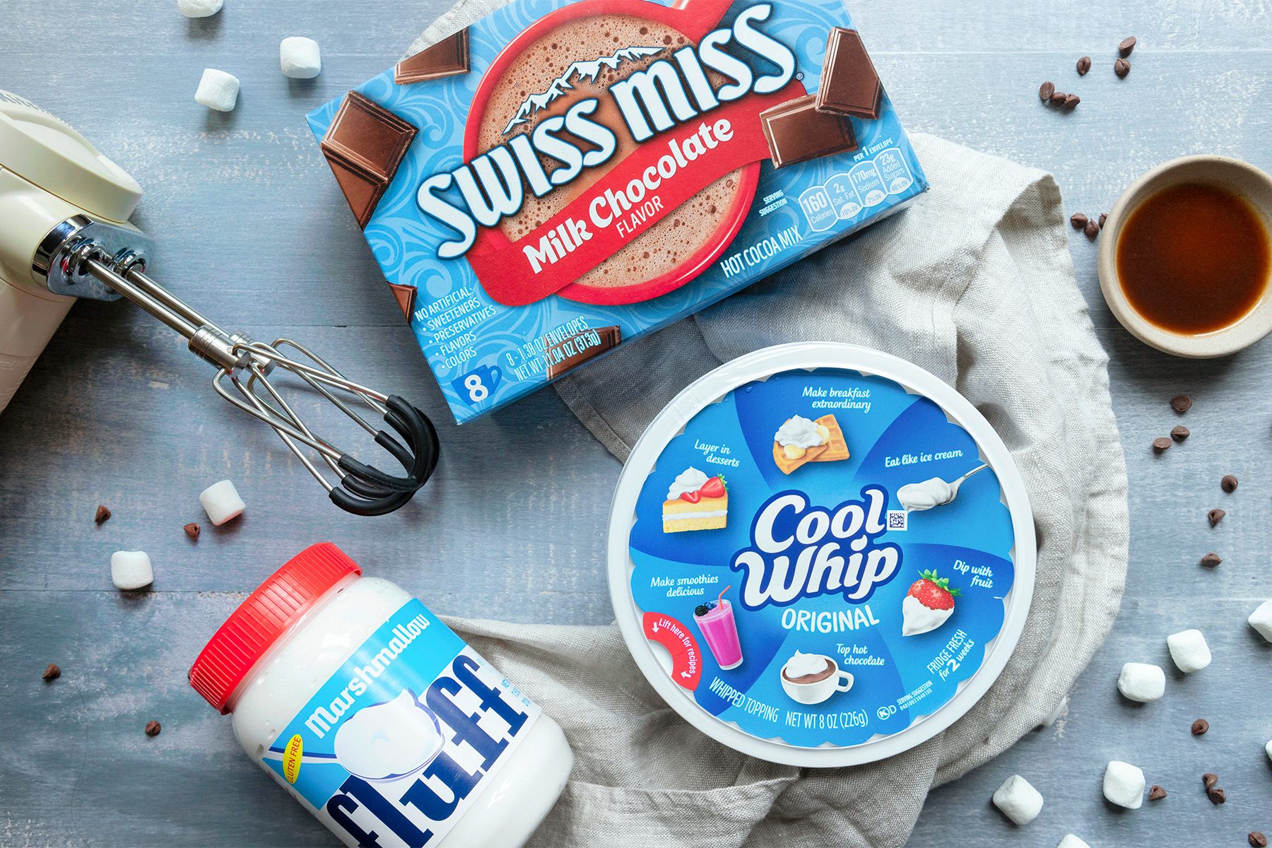A flat lay photo of a box of Swiss Miss milk chocolate, a tub of Cool Whip, and a jar of Fluff marshmallow spread. Nearby are a mixer, small marshmallows, chocolate chips, and a cup of brown liquid on a light grey surface.