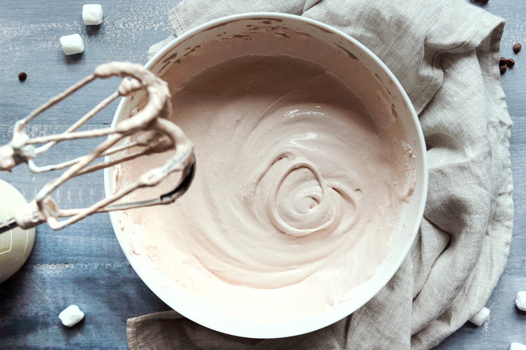 A bowl of creamy, whipped mixture with a hand mixer resting on the side. The bowl is on a wooden surface, partially covered by a beige cloth. Small marshmallows and chocolate chips are scattered around.