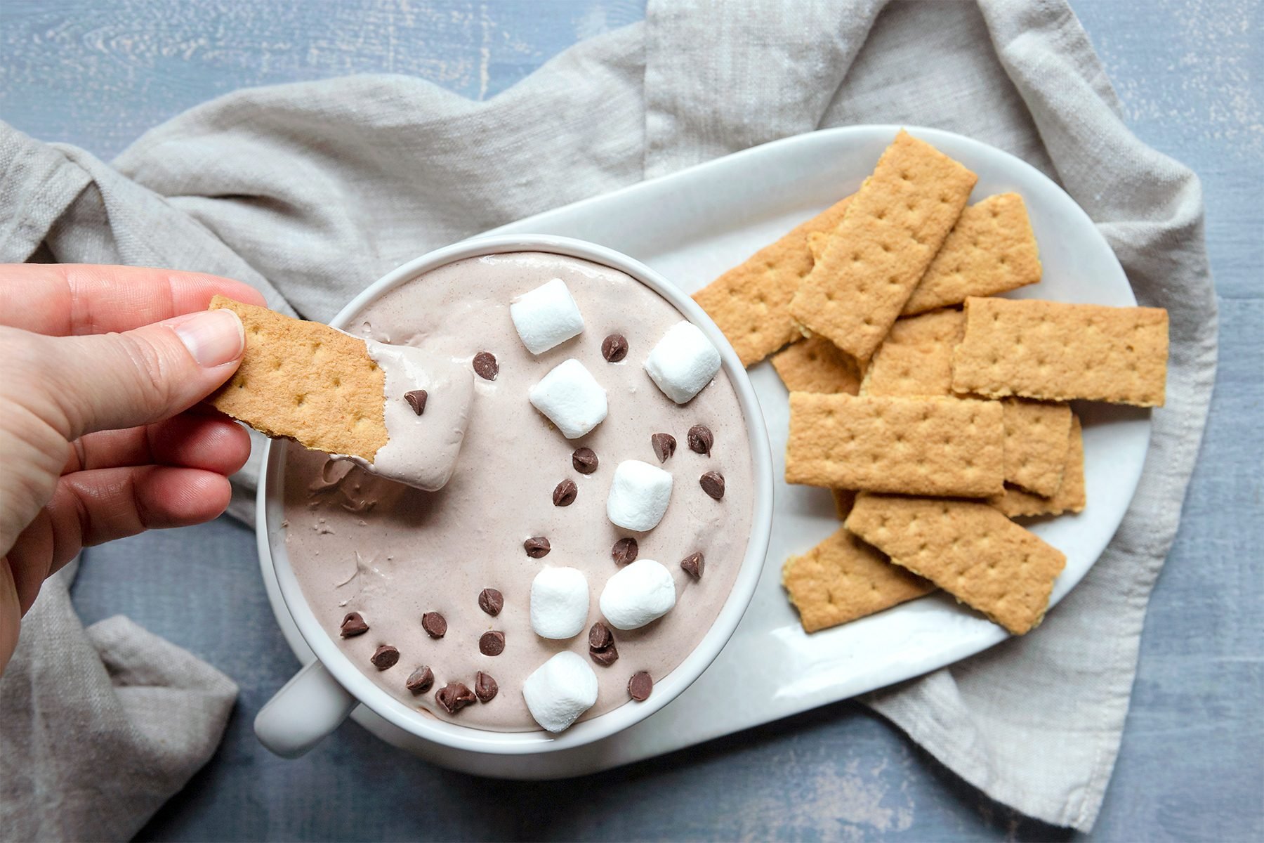 A person dips a graham cracker into a bowl of chocolate dip, topped with marshmallows and chocolate chips. The bowl is on a white platter with more graham crackers, set on a textured gray surface with a beige cloth.