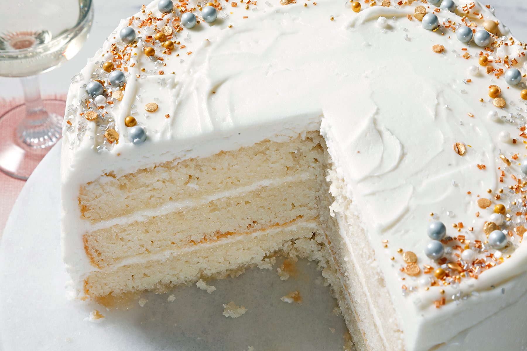 A three-layer vanilla cake with white frosting on a marble surface. The top is decorated with silver balls and golden sprinkles. A slice is missing from the cake. A glass of white wine is partially visible in the background.