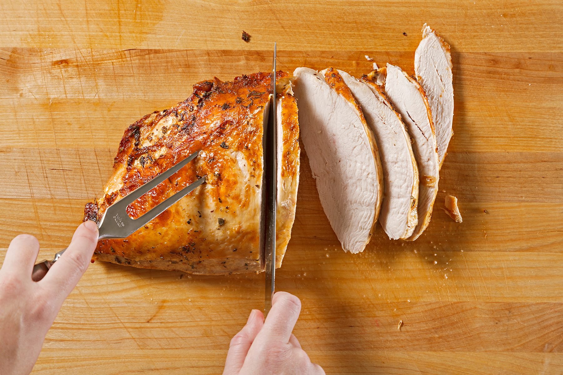 A person slices a cooked, golden-brown piece of turkey breast on a wooden cutting board, revealing several juicy, evenly cut slices. A fork secures the meat while a knife carves through it.