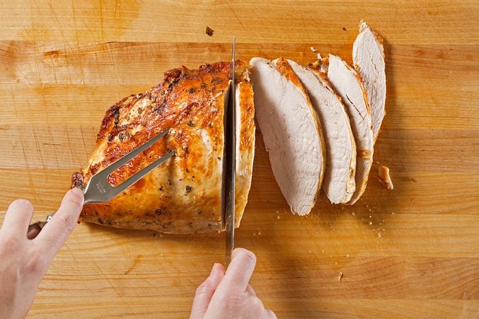 A person slices a cooked, golden-brown piece of turkey breast on a wooden cutting board, revealing several juicy, evenly cut slices. A fork secures the meat while a knife carves through it.