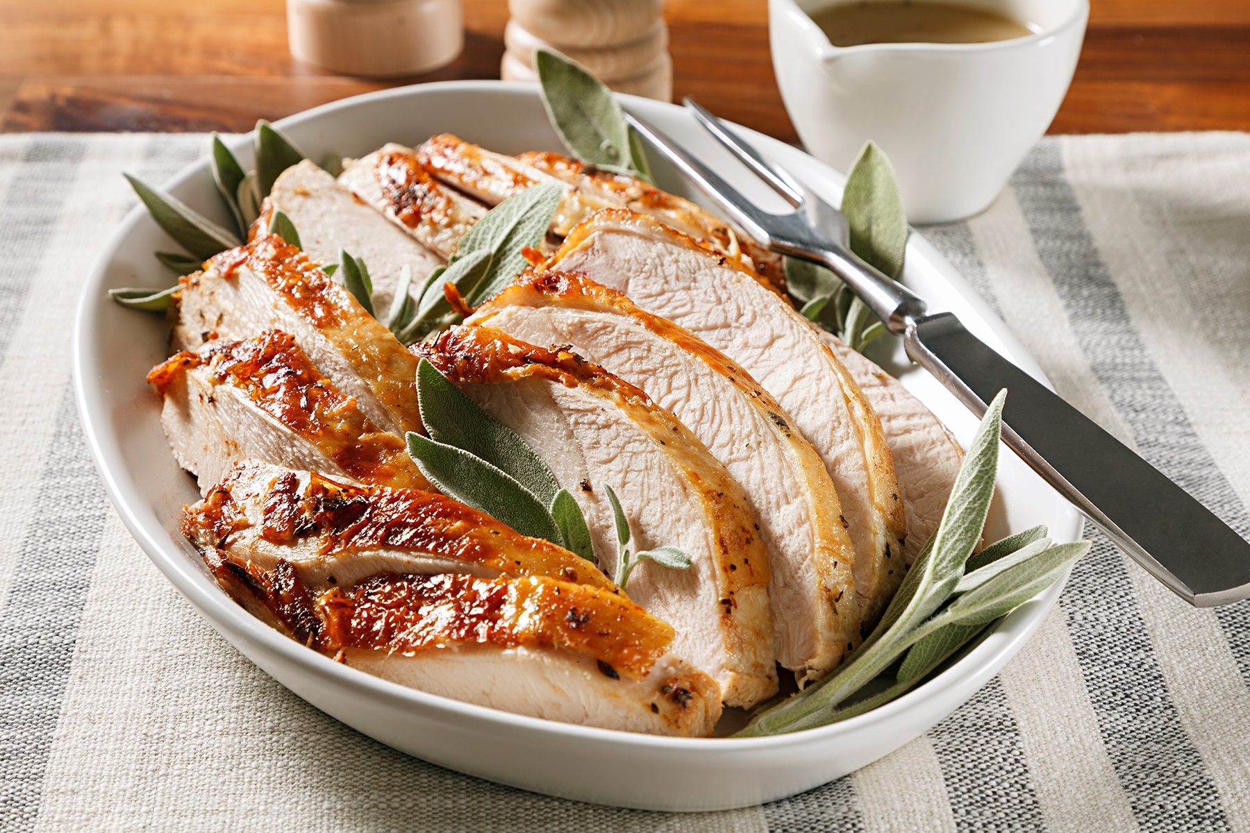 A white dish with sliced roasted turkey breast garnished with fresh sage leaves. A carving fork and knife rest on the side. A small gravy boat is visible in the background. The setting is atop a striped tablecloth.