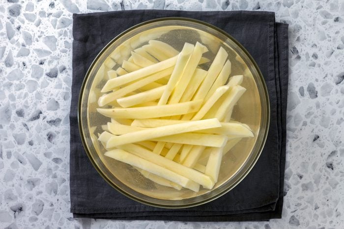 Potato strips covered with water in a medium bowl on kitchen cloth