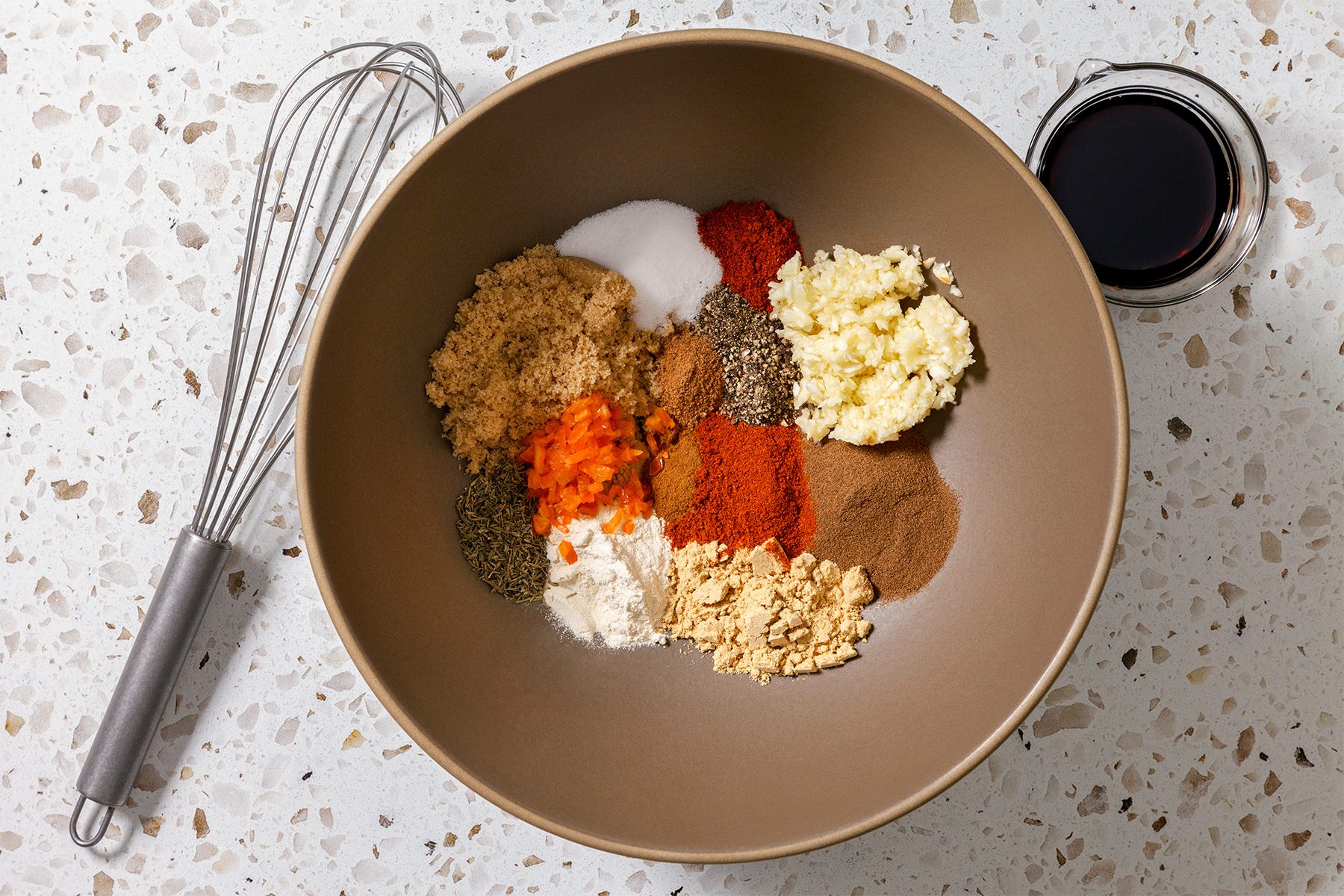 overhead shot of a brown bowl on a white countertop with terrazzo flecks; in the bowl are various spices and ingredients for cooking, next to the bowl is a whisk and a small glass bowl with dark liquid, likely soy sauce