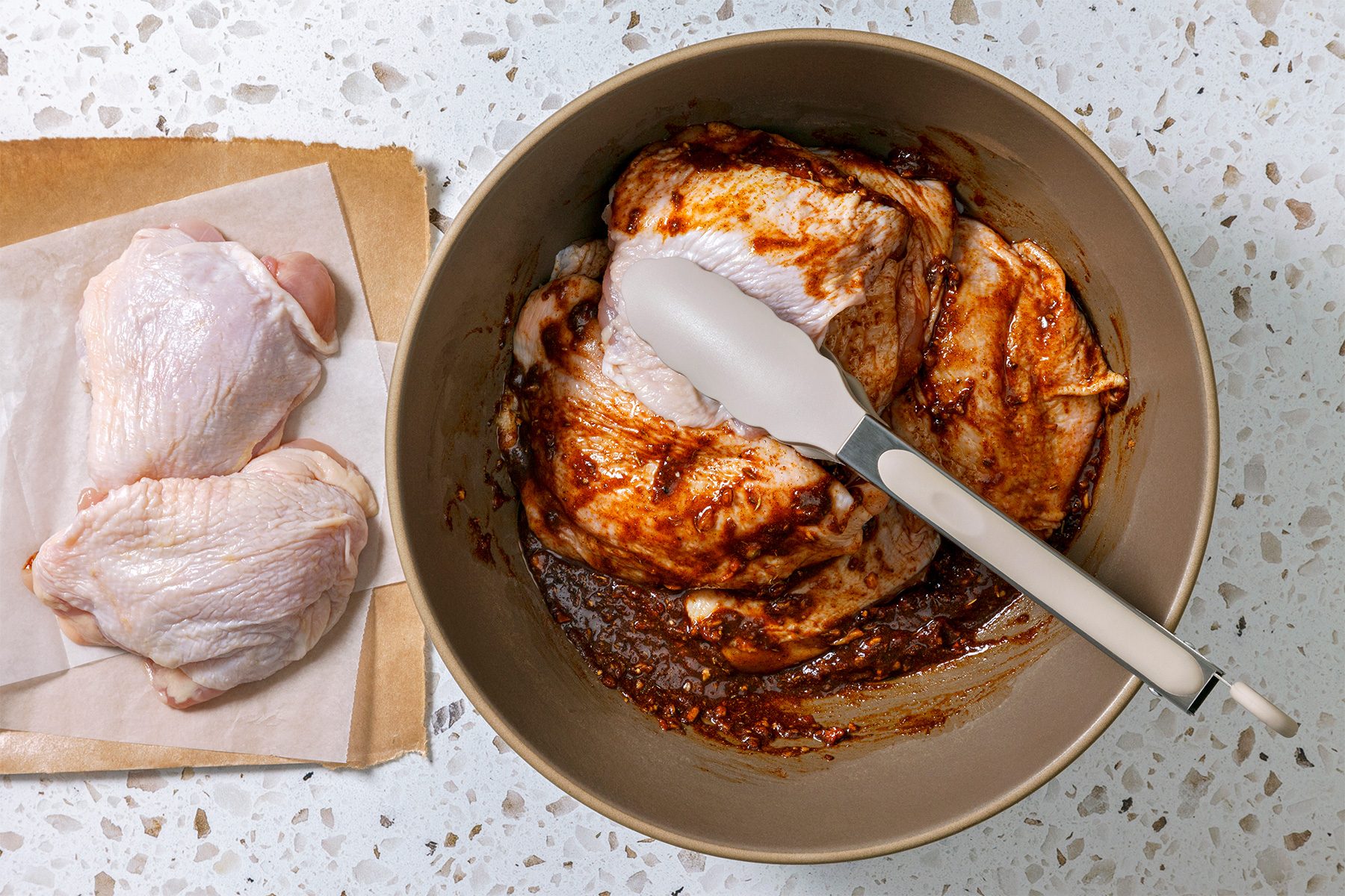 overhead shot of two raw chicken thighs on a piece of parchment paper; they are next to a bowl that contains several chicken thighs coated in a reddish brown marinade, a white spatula with a silver handle is lying over top of the chicken thighs in the bowl; the chicken in the bowl is covered in a thick marinade; the bowl is brown and is sitting on a speckled countertop
