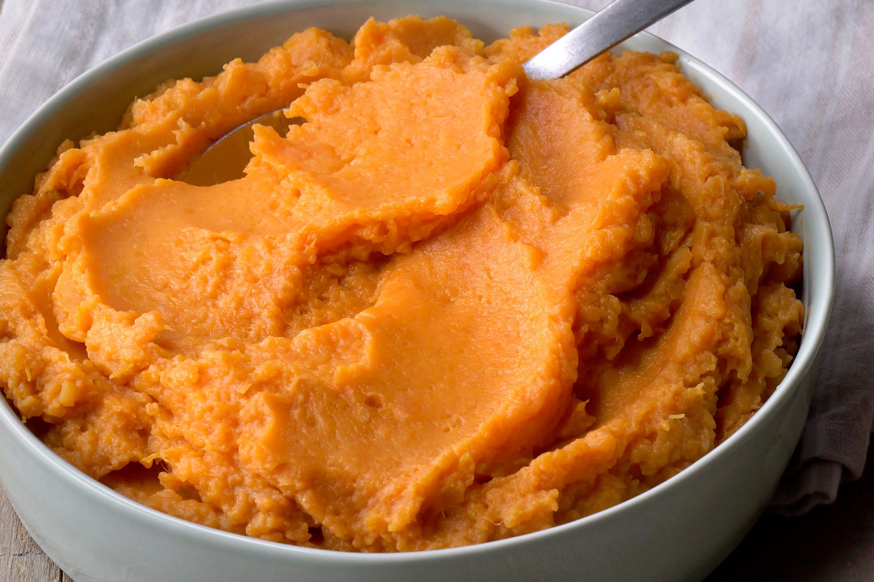 closeup shot of a bowl of mashed sweet potatoes, with a spoon resting on top; the sweet potatoes are a vibrant orange color, and the bowl is a pale blue-green;