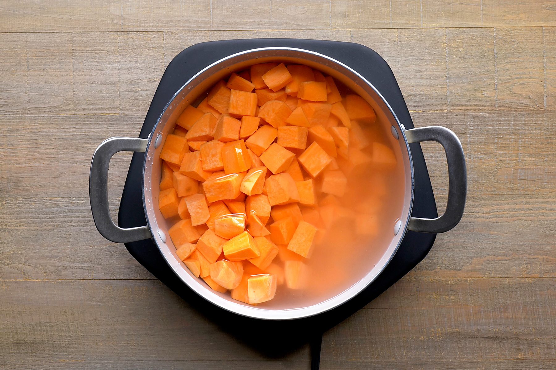 overhead shot of a pot of cubed sweet potatoes submerged in water on a wooden surface; the pot is made of stainless steel with black handles and is sitting on a black heat-resistant pad