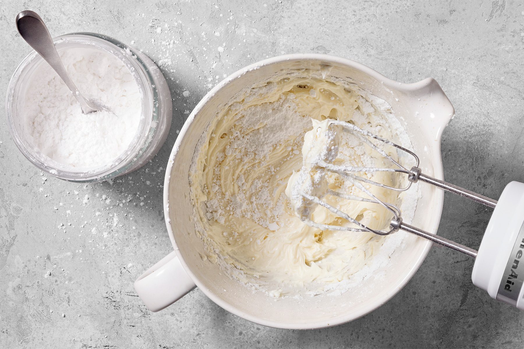 overhead shot of a white bowl containing a mixture of frosting and powdered sugar, with a silver electric mixer whisk inside the bowl; the whisk is connected to a white electric mixer, a glass jar filled with powdered sugar with a silver spoon inside is partially visible on the left side of the image; the bowl and jar are sitting on a gray countertop, with powdered sugar scattered around the bowl and jar