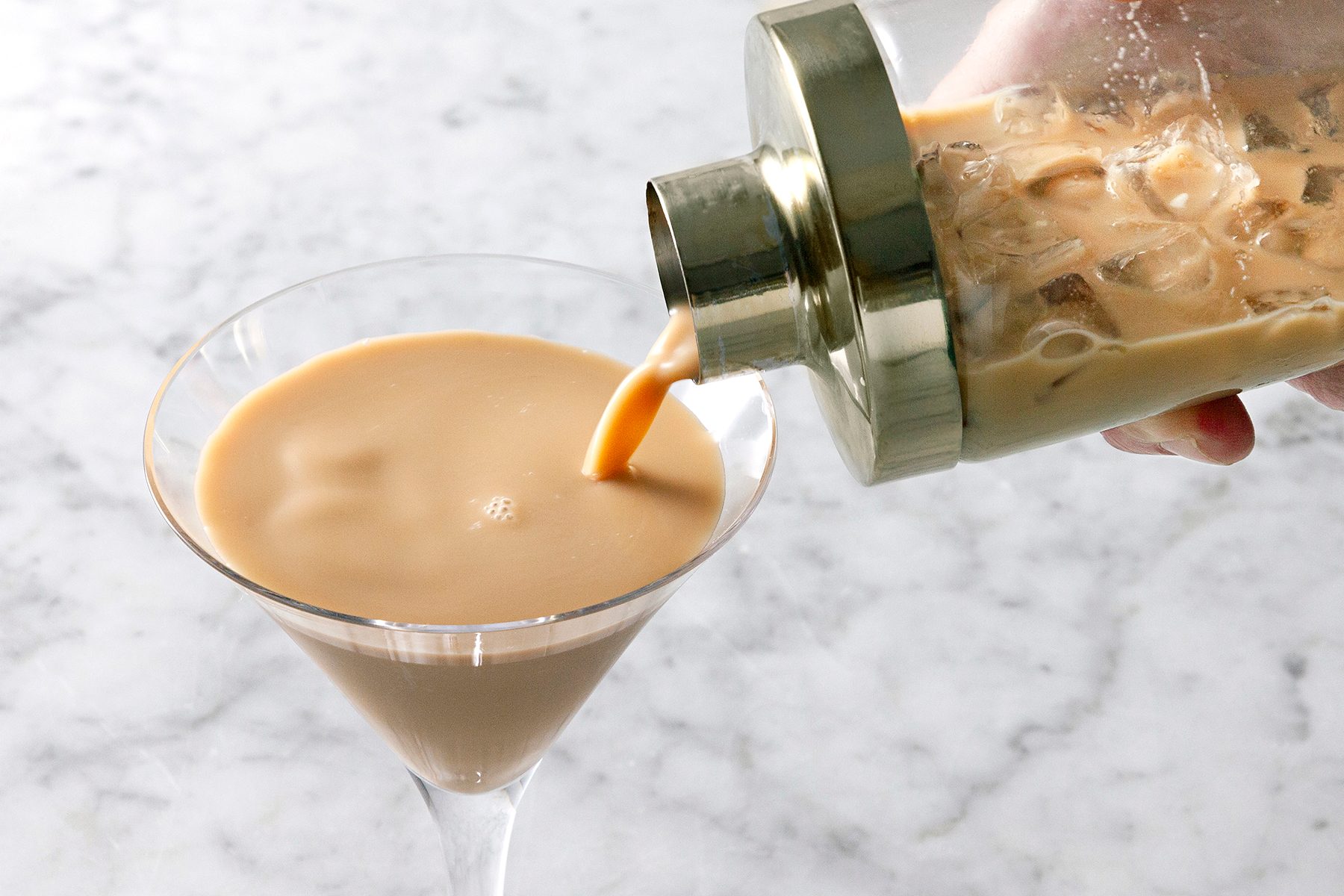 A hand pours a creamy, light brown cocktail from a shaker into a martini glass on a marble countertop.