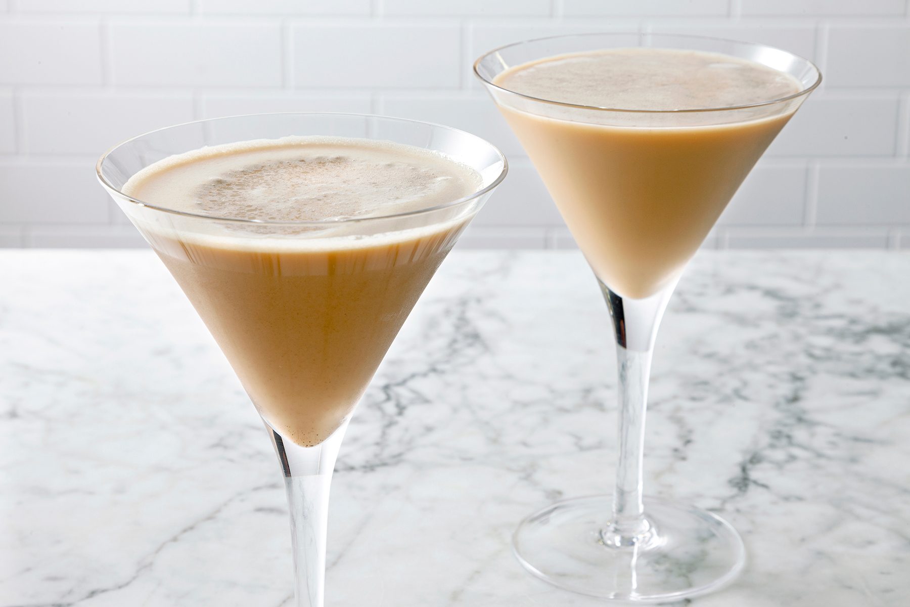 Two martini glasses filled with a creamy beige cocktail are placed on a white marble surface. The drinks have a light frothy top, and the background features a white tiled wall.