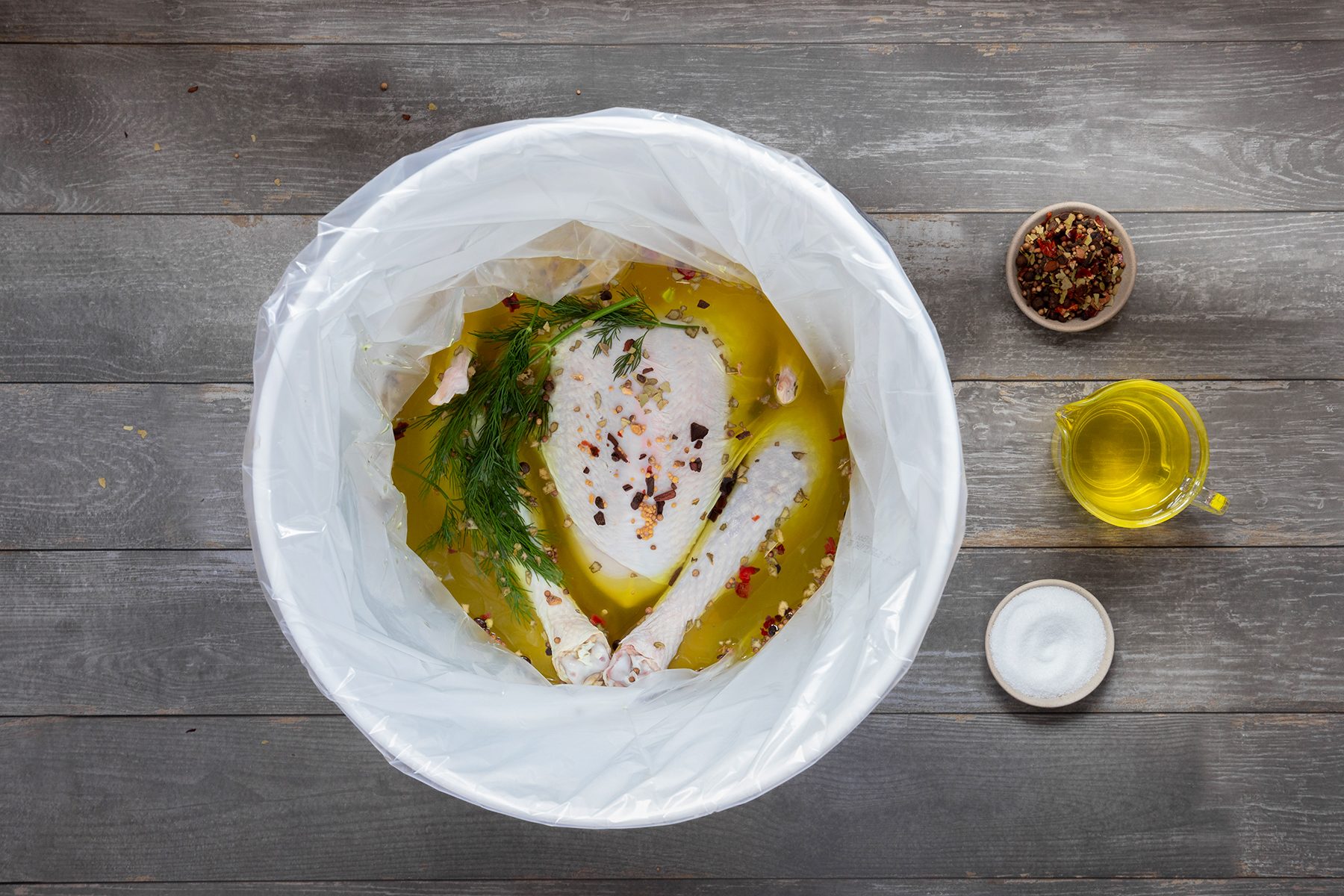 Raw turkey breast and drumsticks marinating in a container with oil, herbs, and spices. A small bowl of mixed spices, a bowl of olive oil, and a bowl of salt are placed beside the container on a wooden surface.