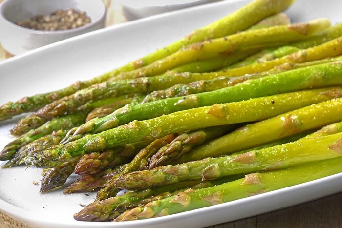 A platter of roasted asparagus is served on a white dish. The spears are drizzled with oil and lightly seasoned with pepper and salt. Small bowls of peppercorns are visible in the background.