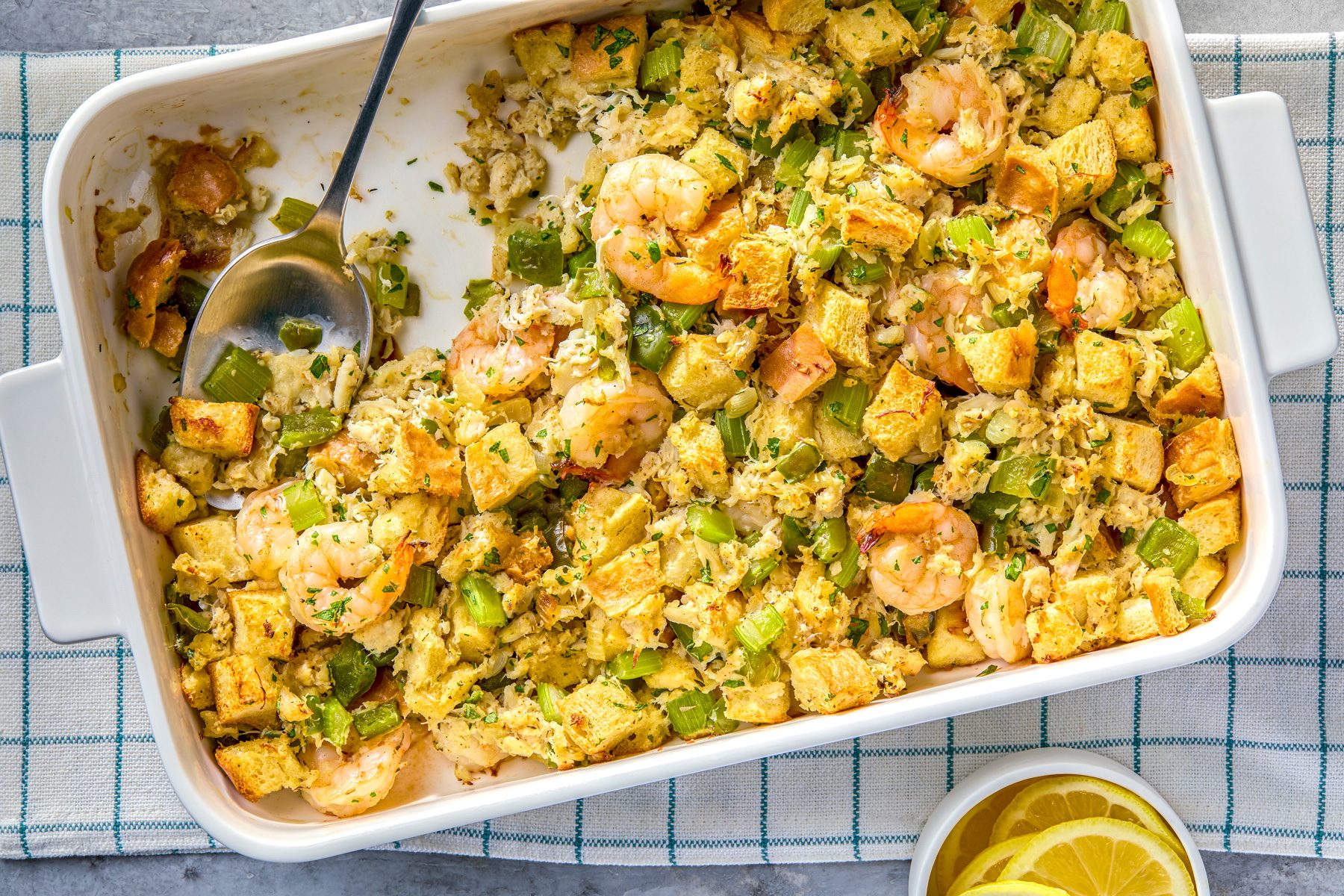 overhead shot of a white baking dish filled with a seafood stuffing is sitting on top of a checkered kitchen towel; a metal spoon is lying partially submerged in the stuffing, a small white bowl filled with lemon slices is sitting on the towel to the right of the baking dish