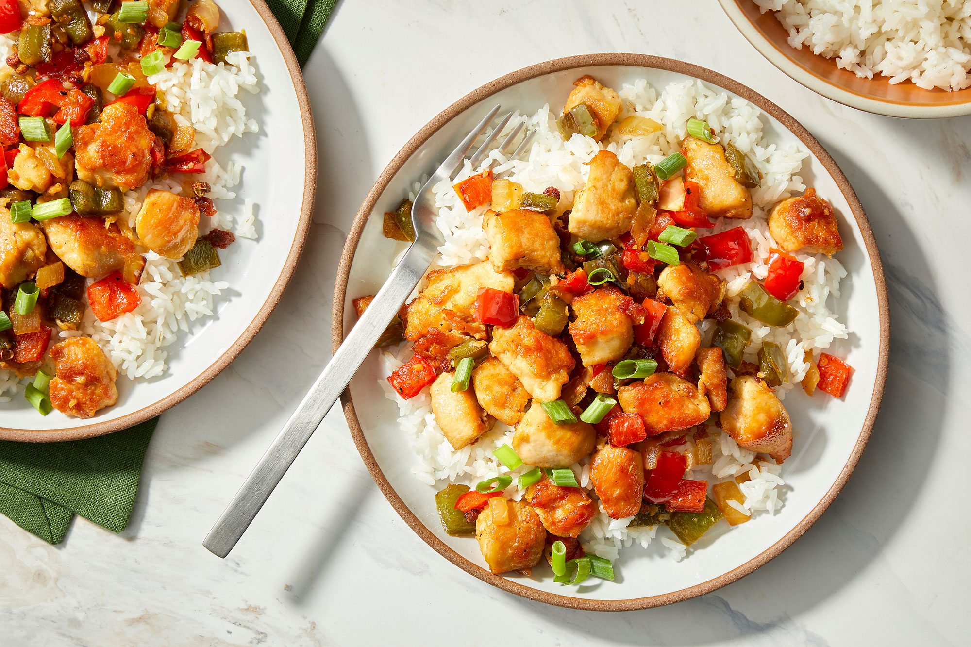 A plate of food that contains Szechuan chicken, rice, green peppers, red peppers and green onions, all of which are cooked in a sauce