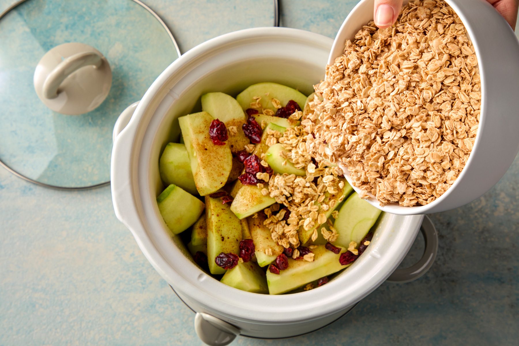overhead shot of a white bowl with oats is being poured into a white crock pot that contains sliced apples and cranberries; the crock pot is on a blue and white speckled surface , with a white lid to the side , there is a hand holding the bowl of oats, ready to pour them into the crock pot