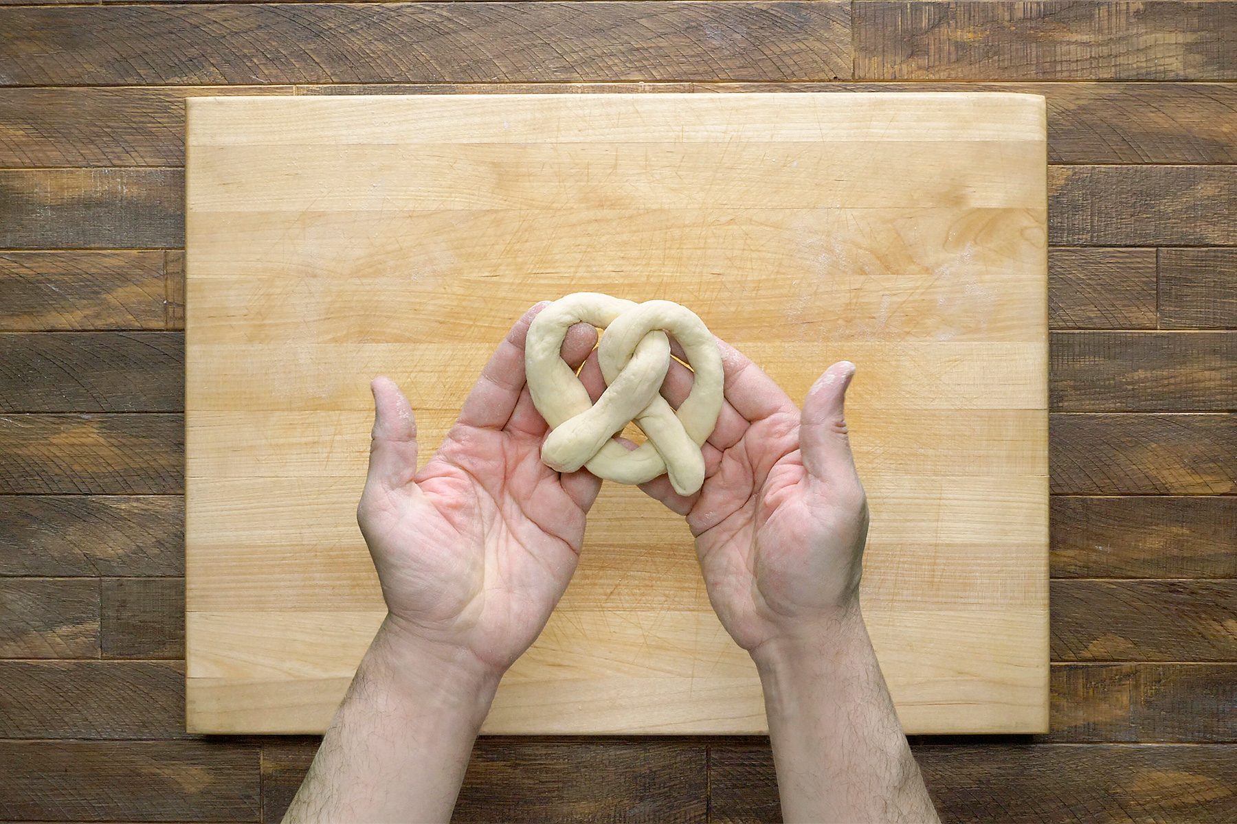 Two hands holding a raw pretzel dough in the shape of a twist above a wooden cutting board on a wooden surface.